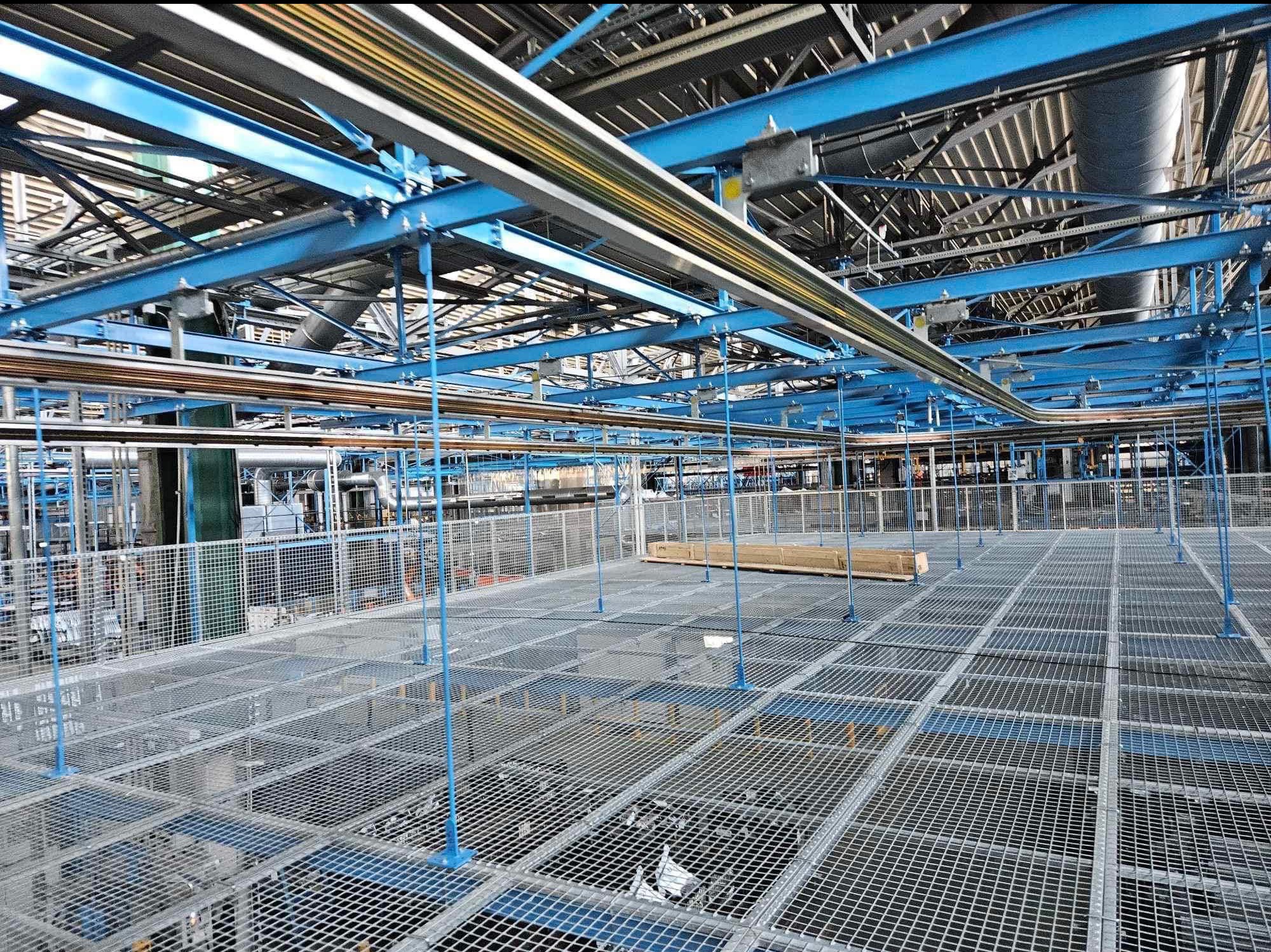 Rozległa hala przemysłowa z metalową kratownicą podłogową, niebieskimi konstrukcjami wsporczymi i rzędami przewodów elektrycznych pod sufitem.