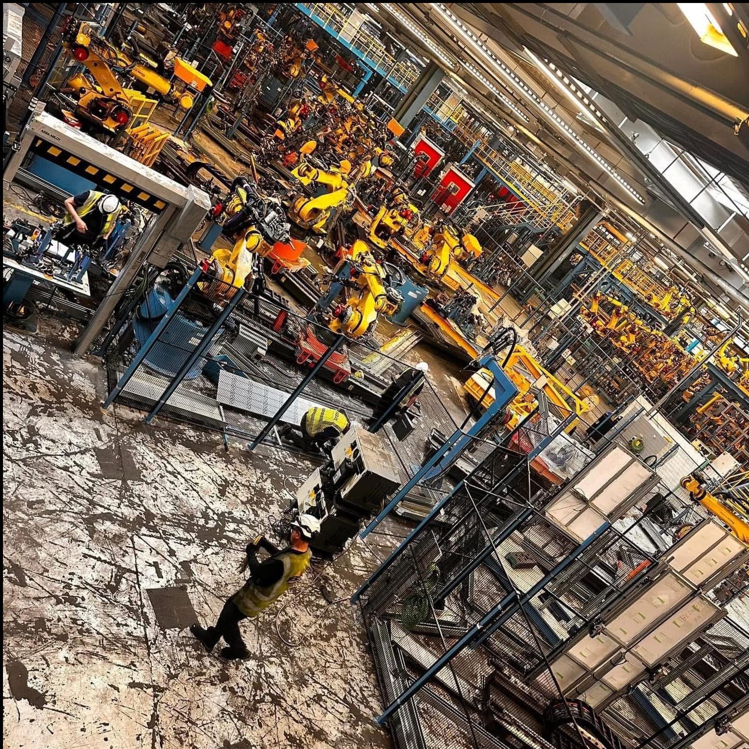 Rozległa hala fabryczna z licznymi żółtymi robotami przemysłowymi; pracownicy w kamizelkach i kaskach przy stanowiskach. Widok z góry, perspektywa zniekształcona.