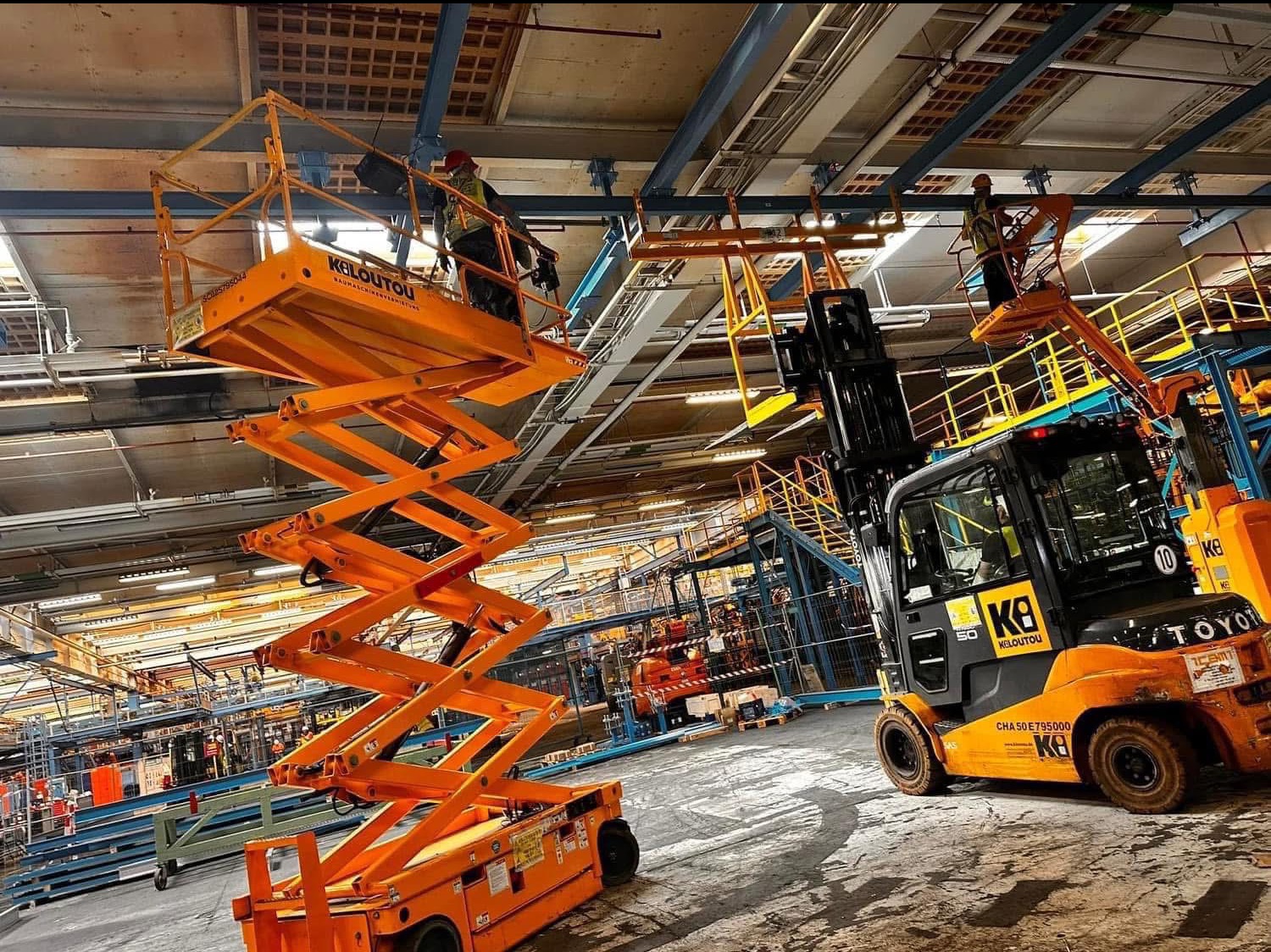 Praca w hali: elektryk na podnośniku montuje oświetlenie pod sufitem. W tle widoczny wózek widłowy i inne podnośniki. Drobne prace na wysokości.