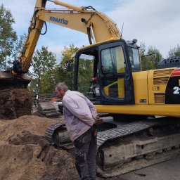 DT Advenced SP.ZO.O. - Operator obserwuje żółtą koparkę Komatsu PC 210 LC podczas prac ziemnych, z widoczną stertą ziemi w łyżce i piasku na ziemi.