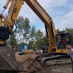 DT Advenced SP.ZO.O. - Żółta koparka Komatsu na placu budowy, operator w kabinie, obok pracownik w roboczym ubraniu i grupie osób w tle.