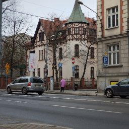 DT Advenced SP.ZO.O. - Kamienica z wieżyczką w stylu pruskim, widoczna z ulicy z przejeżdżającymi samochodami i pieszą w różowym płaszczu.