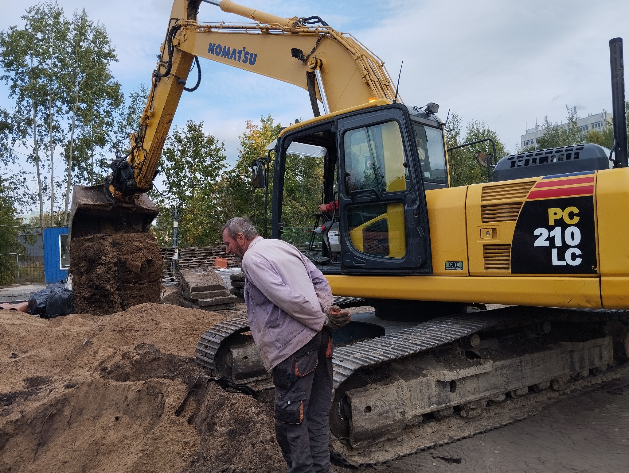 Operator obserwuje żółtą koparkę Komatsu PC 210 LC podczas prac ziemnych, z widoczną stertą ziemi w łyżce i piasku na ziemi.