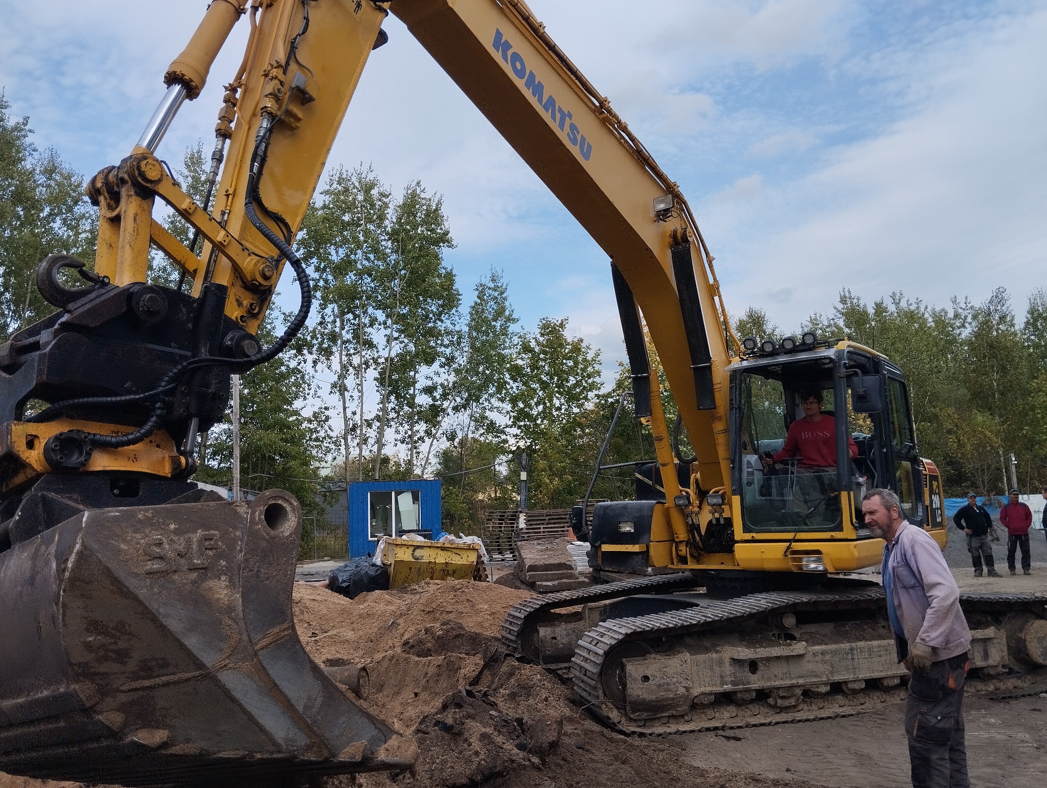 Żółta koparka Komatsu na placu budowy, operator w kabinie, obok pracownik w roboczym ubraniu i grupie osób w tle.