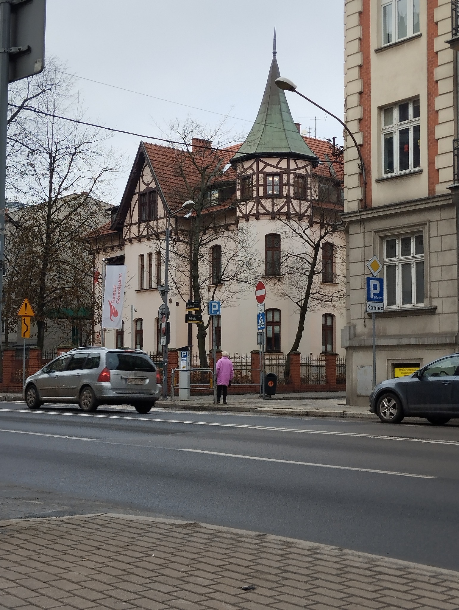 Kamienica z wieżyczką w stylu pruskim, widoczna z ulicy z przejeżdżającymi samochodami i pieszą w różowym płaszczu.