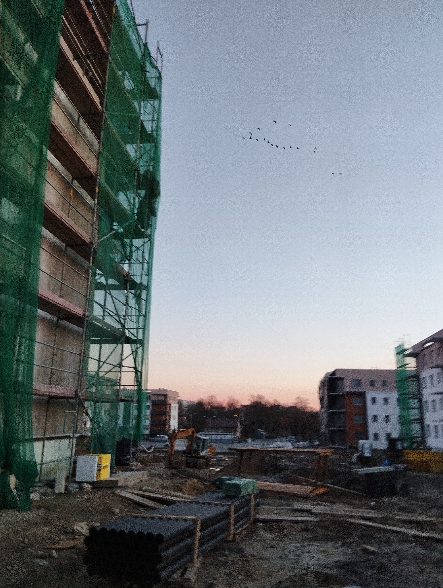 Budynek w trakcie ocieplania, widoczne rusztowania z zieloną siatką ochronną, na pierwszym planie rury izolacyjne, w tle inne budynki i zachodzące słońce oraz klucz ptaków na niebie.