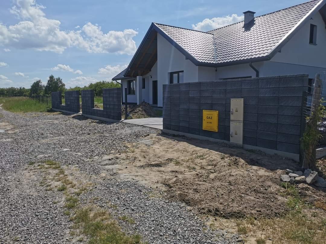 Nowoczesne ogrodzenie modułowe z szarego betonu architektonicznego, z żółtą skrzynką gazową, na tle białego domu z ciemnym dachem i nieba z chmurami.