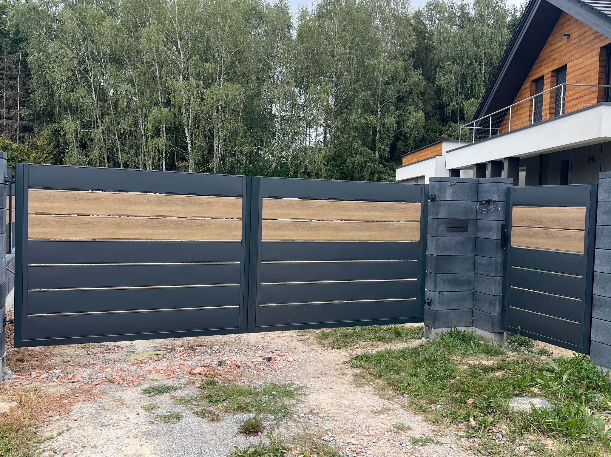 Nowoczesna, przesuwna brama w kolorze antracytowym z poziomymi panelami z drewna kompozytowego, zamontowana na słupkach z szarej kostki betonowej, na tle domu z drewnianą elewacją i zieleni.