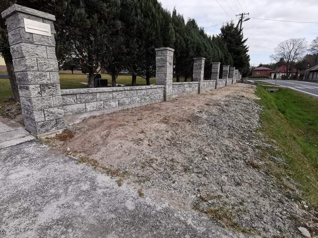 Nowoczesne ogrodzenie z szarej kostki betonowej z filarami, oddzielające posesję od drogi, z widocznymi drzewami iglastymi w tle.