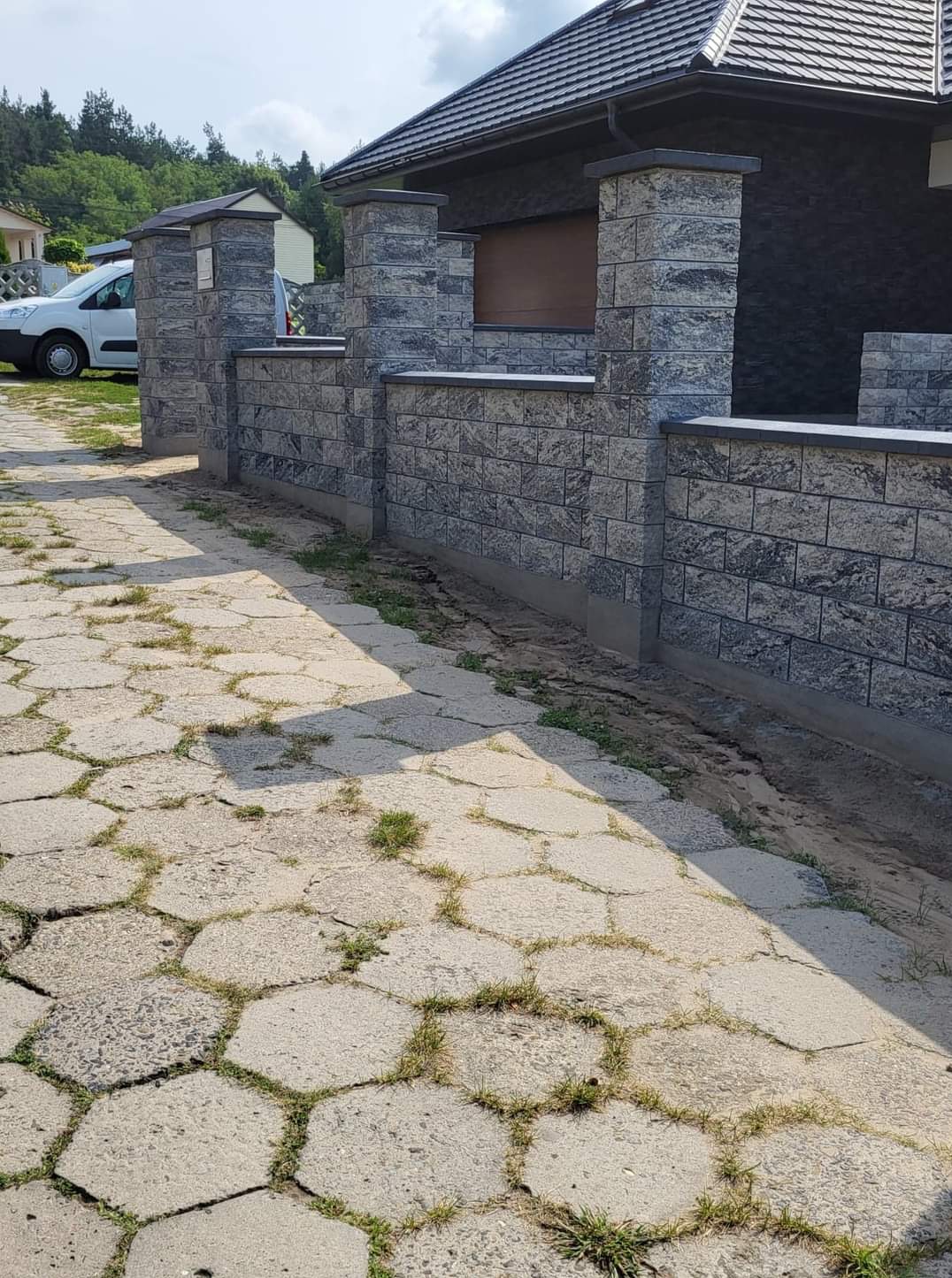 Nowoczesne ogrodzenie z szarej kostki łupanej z elementami słupków, przylegające do budynku z ciemną elewacją, widoczne z poziomu chodnika z betonowych płyt.