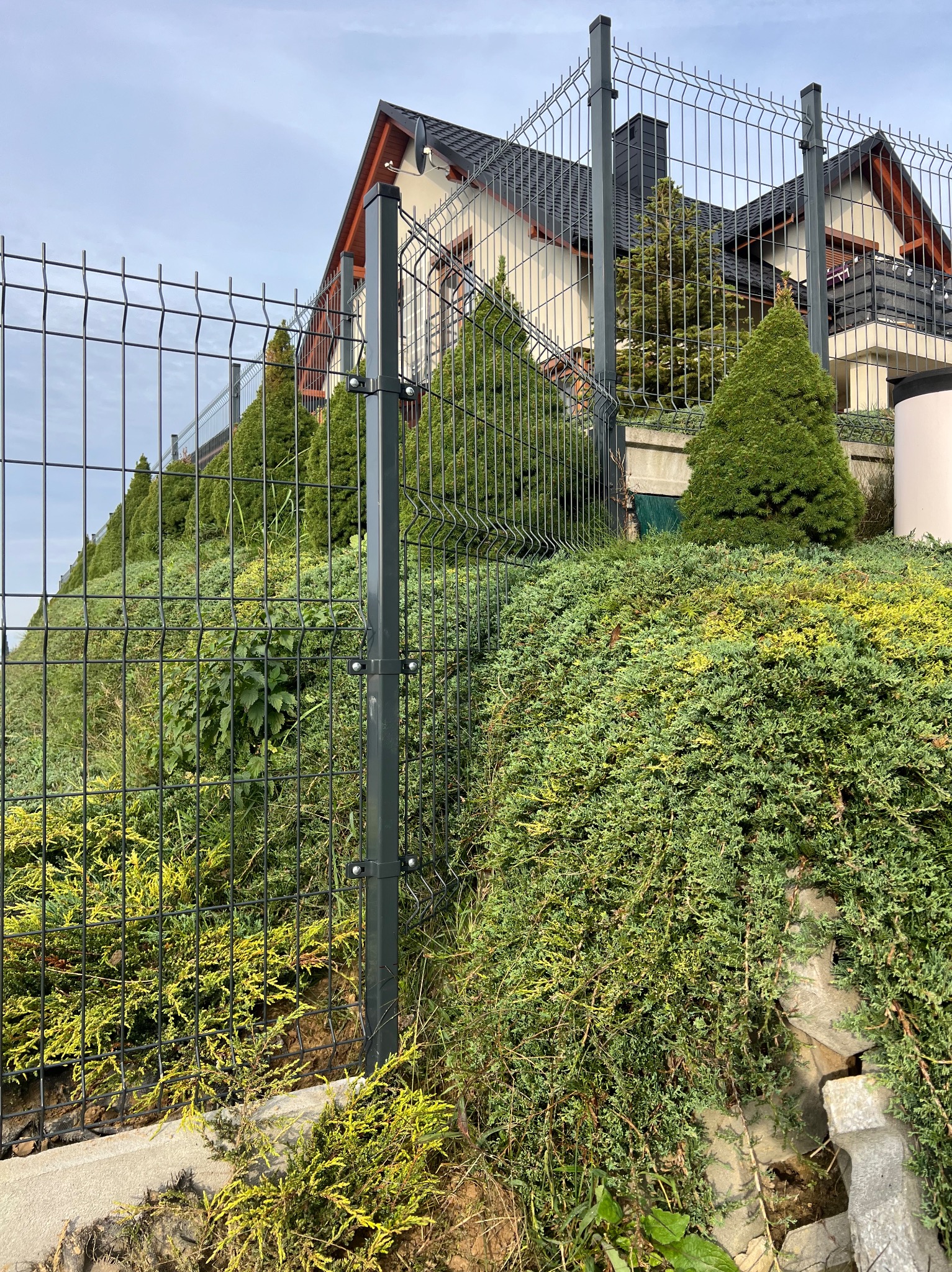 Panelowe ogrodzenie z metalu na nierównym terenie, z widokiem na dom z ciemnym dachem i zielone krzewy. Słupki ogrodzeniowe w kolorze grafitowym.