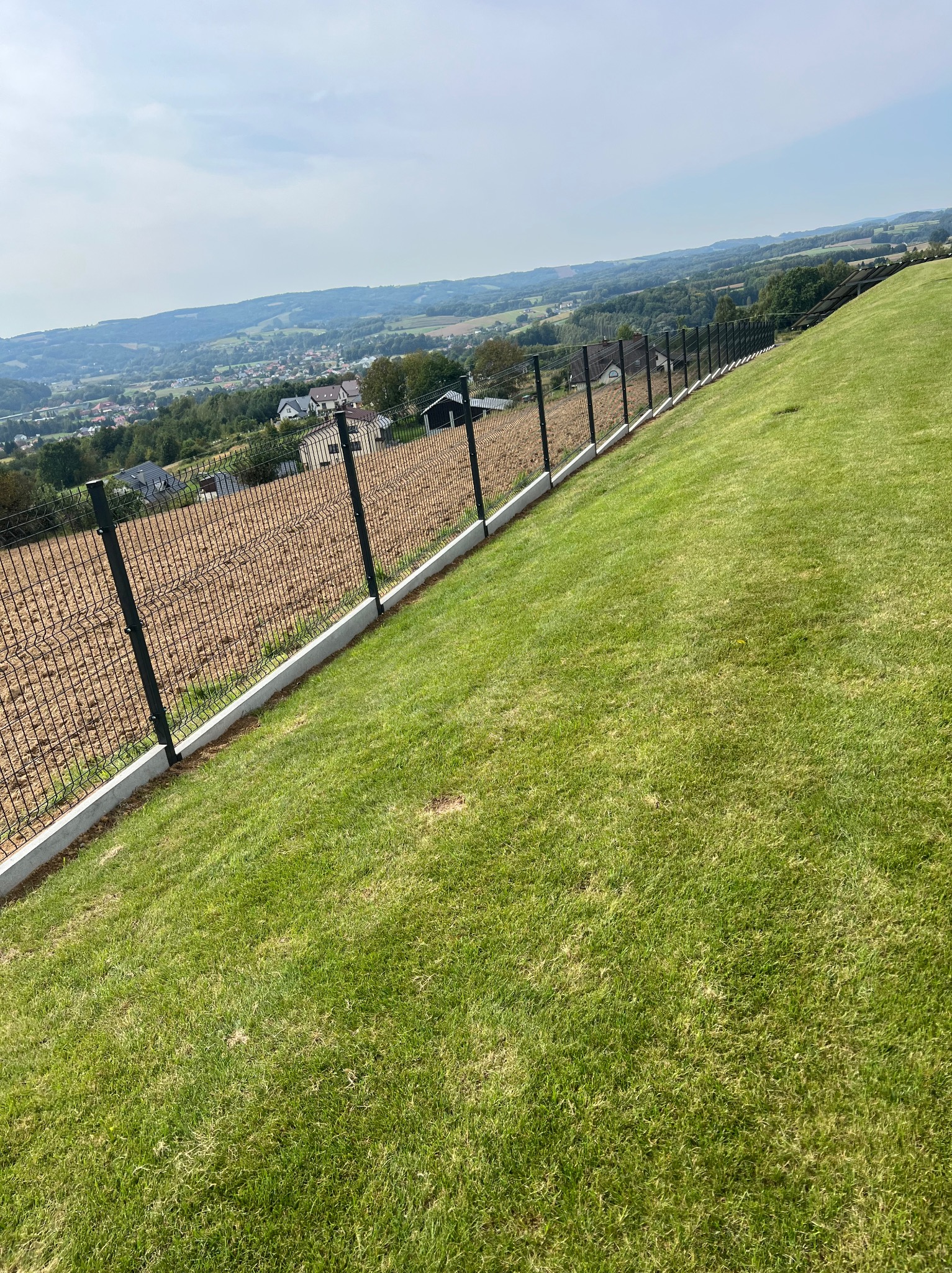 Panelowe ogrodzenie z podmurówką na zboczu, widok na dolinę z domami w oddali. Estetyczne wykończenie terenu.