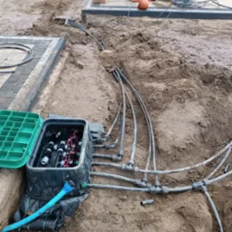 Instalacja podziemnego systemu nawadniania: skrzynka sterująca z otwartą zieloną pokrywą, rury rozprowadzające w wykopie, ziemia wokół.