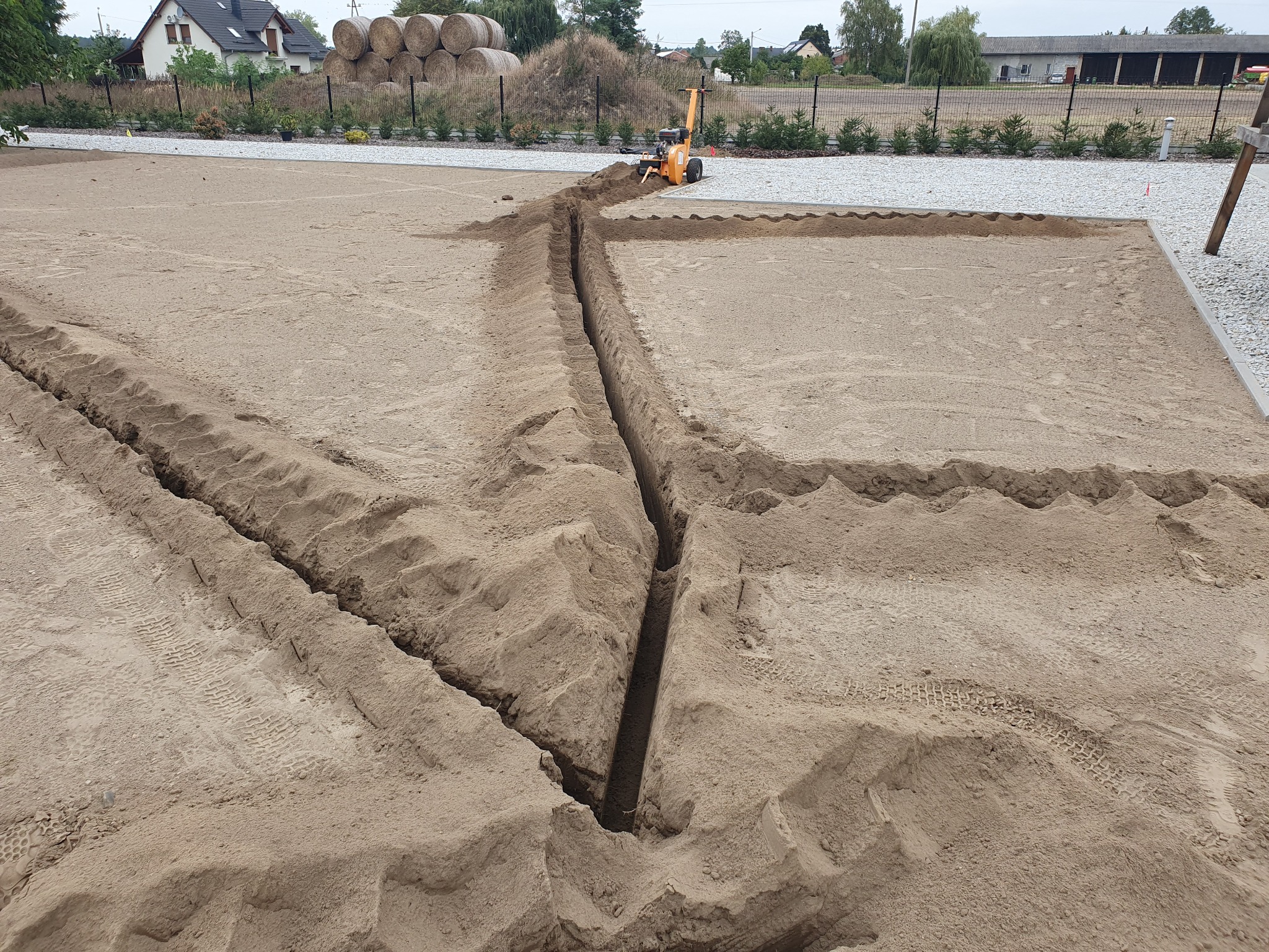 Wykopany rów w piaszczystej ziemi pod instalacje, widoczna mała koparka na tle stogów siana i zabudowań.