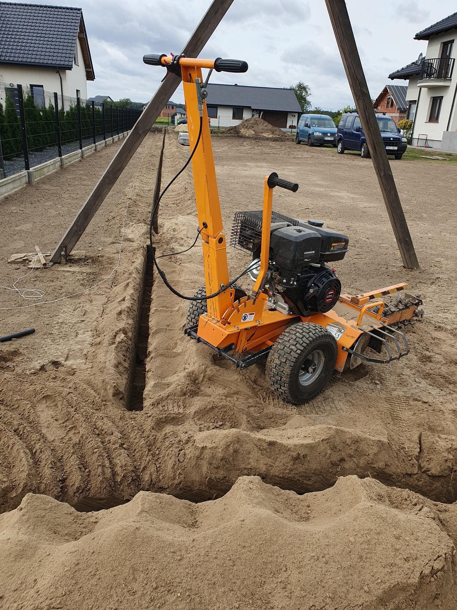 Pomarańczowa glebogryzarka spalinowa do wykopów na piaszczystym terenie, widoczna długa bruzda, domy w tle.