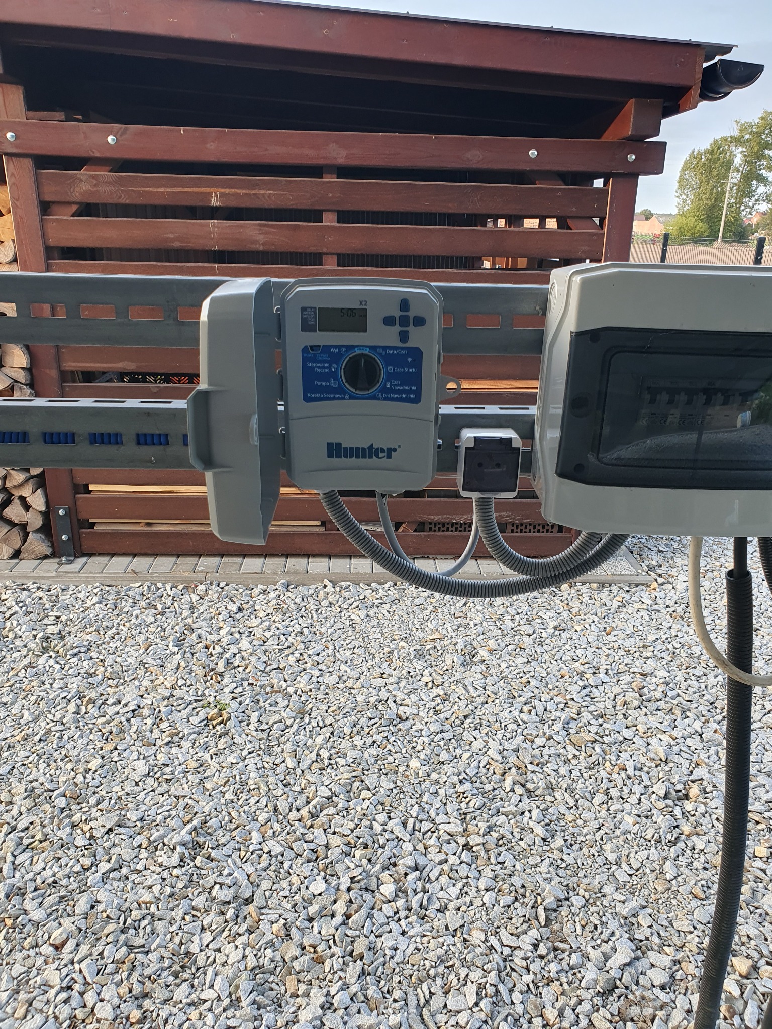 Sterownik systemu nawadniania Hunter X2 zamontowany na metalowej konstrukcji z drewnianym zadaszeniem w tle, widoczne przewody i kostka elektryczna, podłoże wysypane drobnym kamieniem.