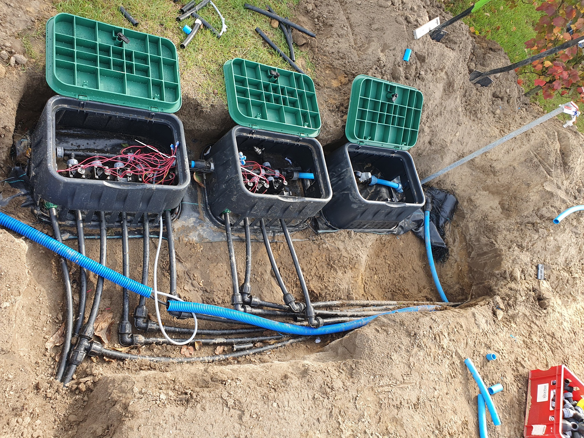 Potrójna instalacja podziemnych zaworów z zielonymi pokrywami w wykopie, widoczne rury i okablowanie, częściowo zasypane ziemią.