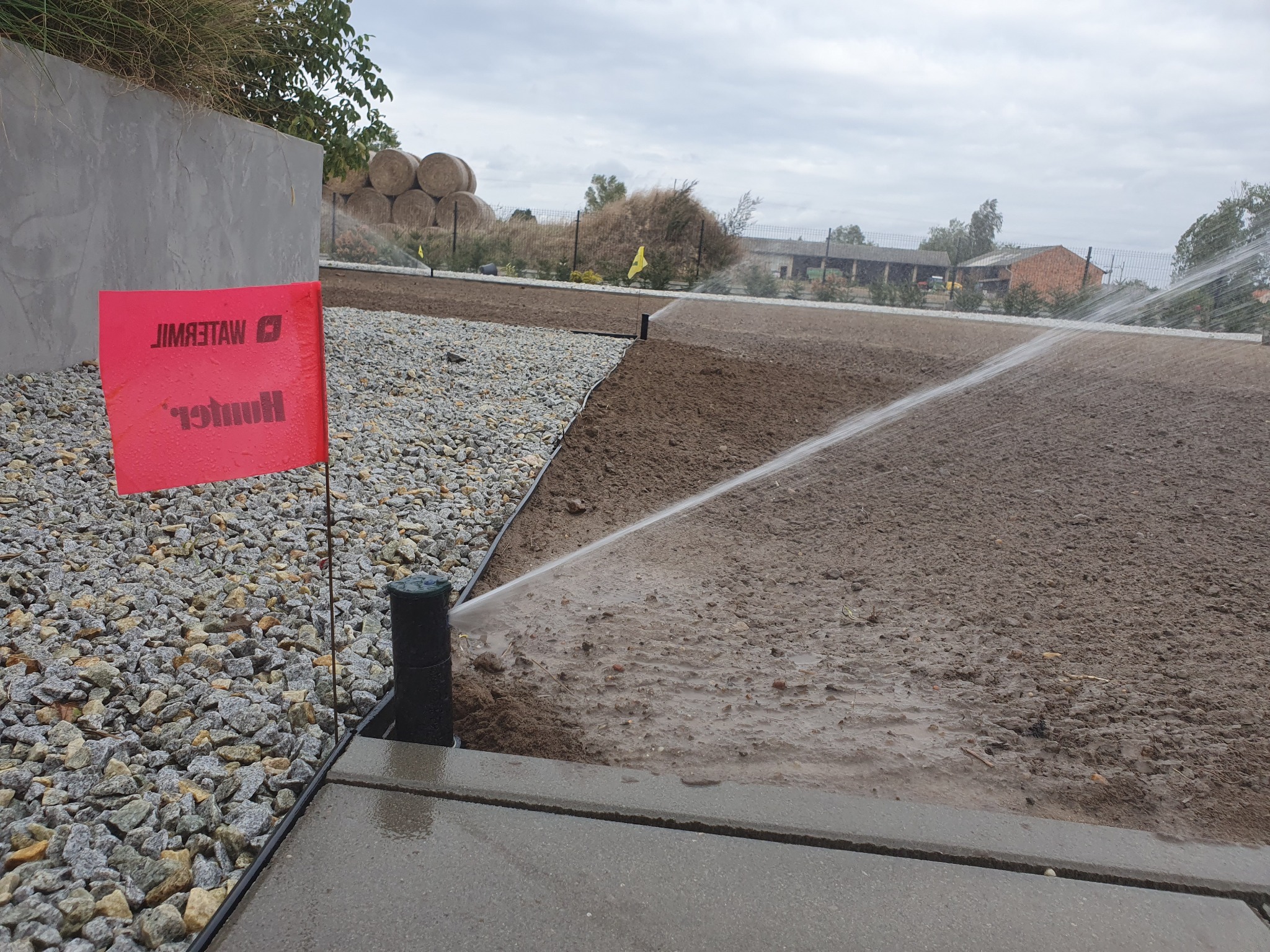 Automatyczny system nawadniania w trakcie pracy, z widocznym czerwonym znacznikiem Hunter, zraszający świeżo przygotowaną glebę na tle sterty bel siana i pochmurnego nieba.