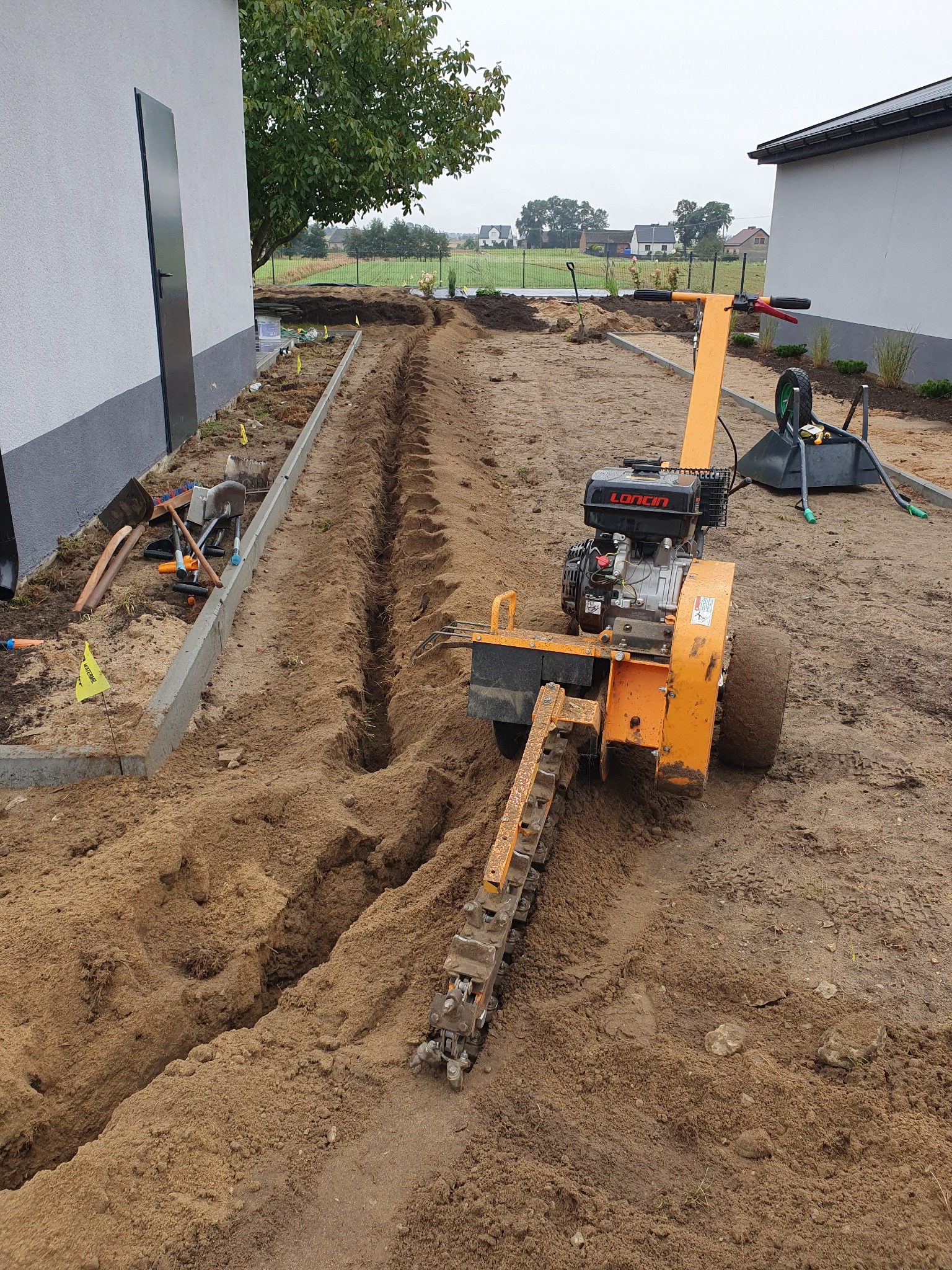 Pomarańczowa glebogryzarka łańcuchowa wykopuje wąski rów w piaszczystej glebie, widoczne narzędzia i taczka w tle.