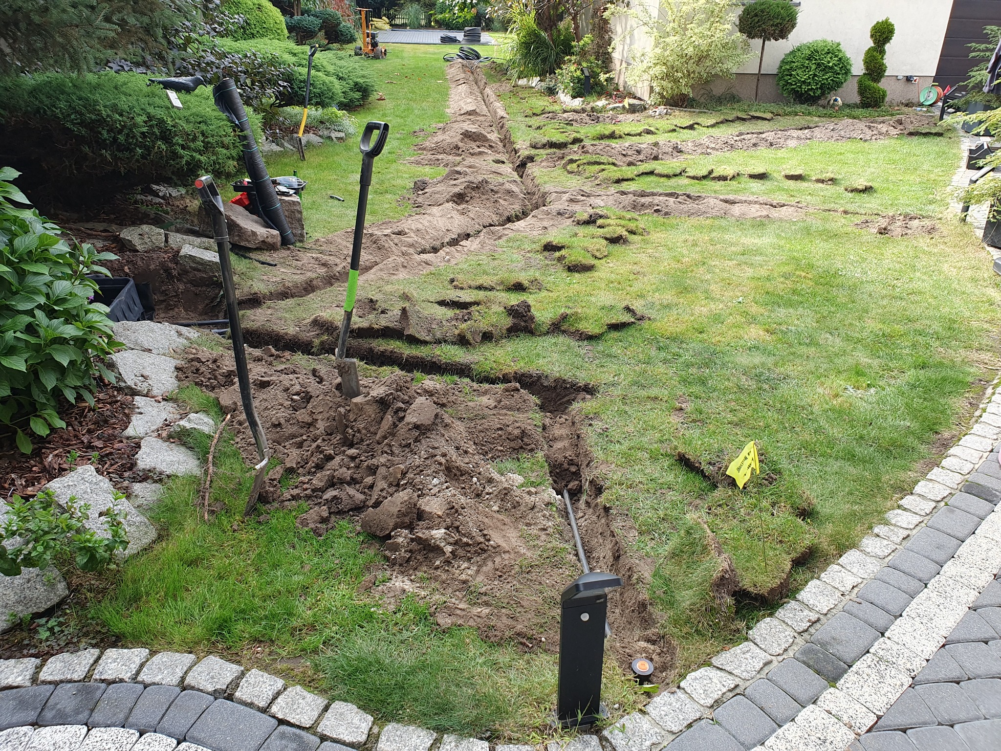 Wykopany wąski rów w trawniku ogrodu, widoczne narzędzia: szpadle i rury, żółta flaga ostrzegawcza, krawężnik z kostki brukowej.