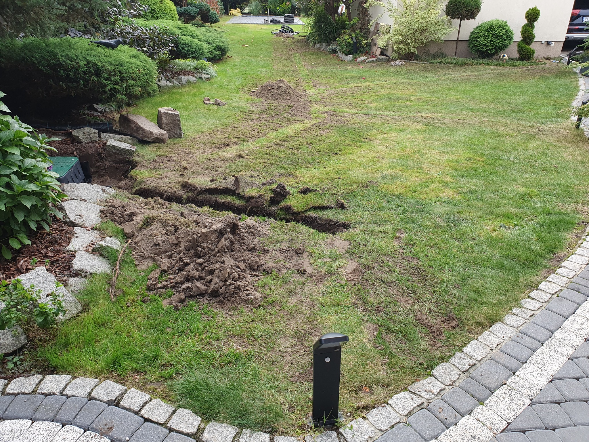 Świeżo wykopane rowy w trawniku, prawdopodobnie pod instalację nawadniającą, z widocznymi stertami ziemi i naruszonym trawnikiem, obok kamienny chodnik i lampa ogrodowa