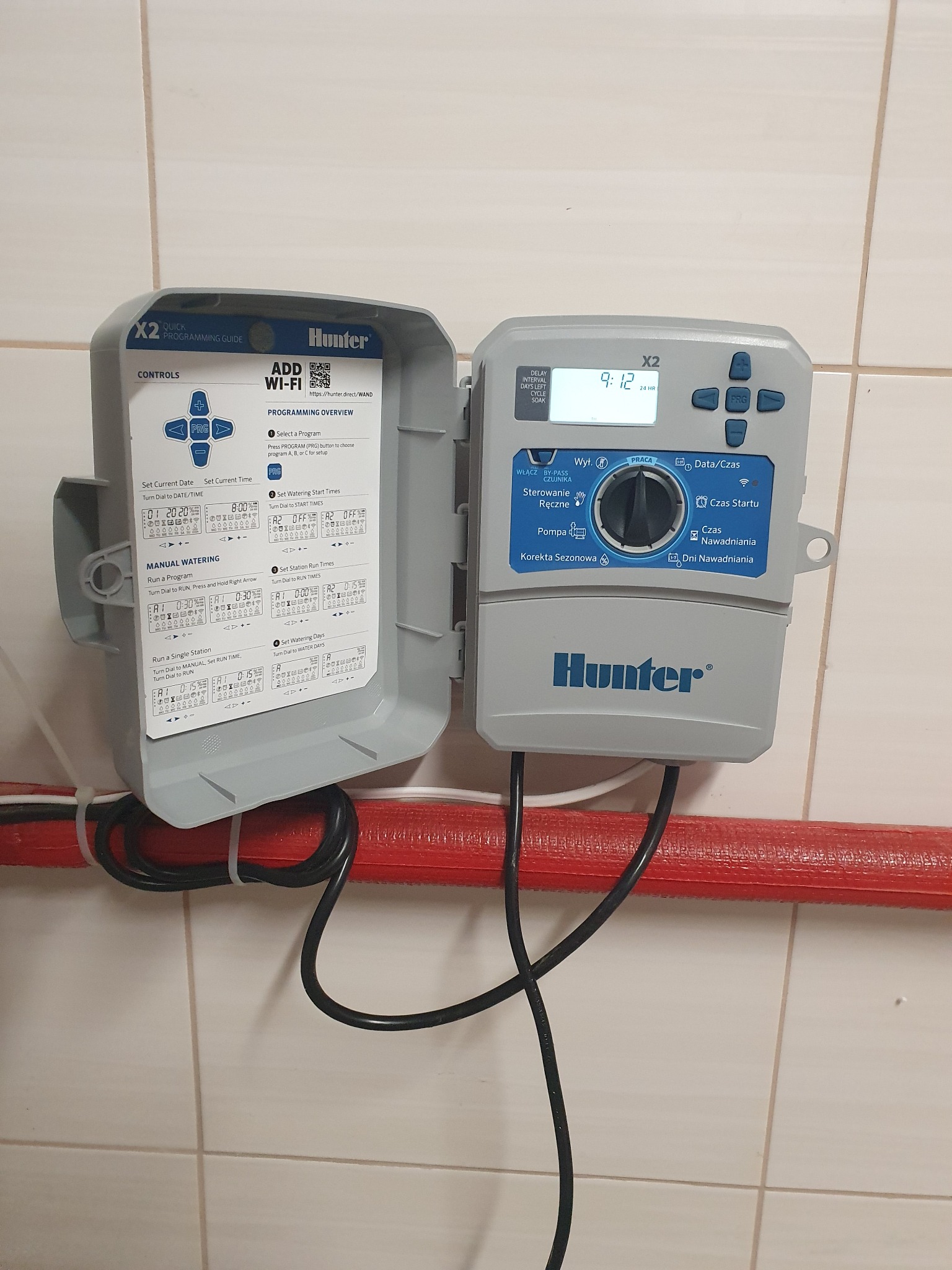 Sterownik nawadniania Hunter X2 z otwartą obudową, widoczny panel sterowania z wyświetlaczem i instrukcją programowania, zamontowany na ścianie z kafelkami, z przewodami przymocowanymi do rury.