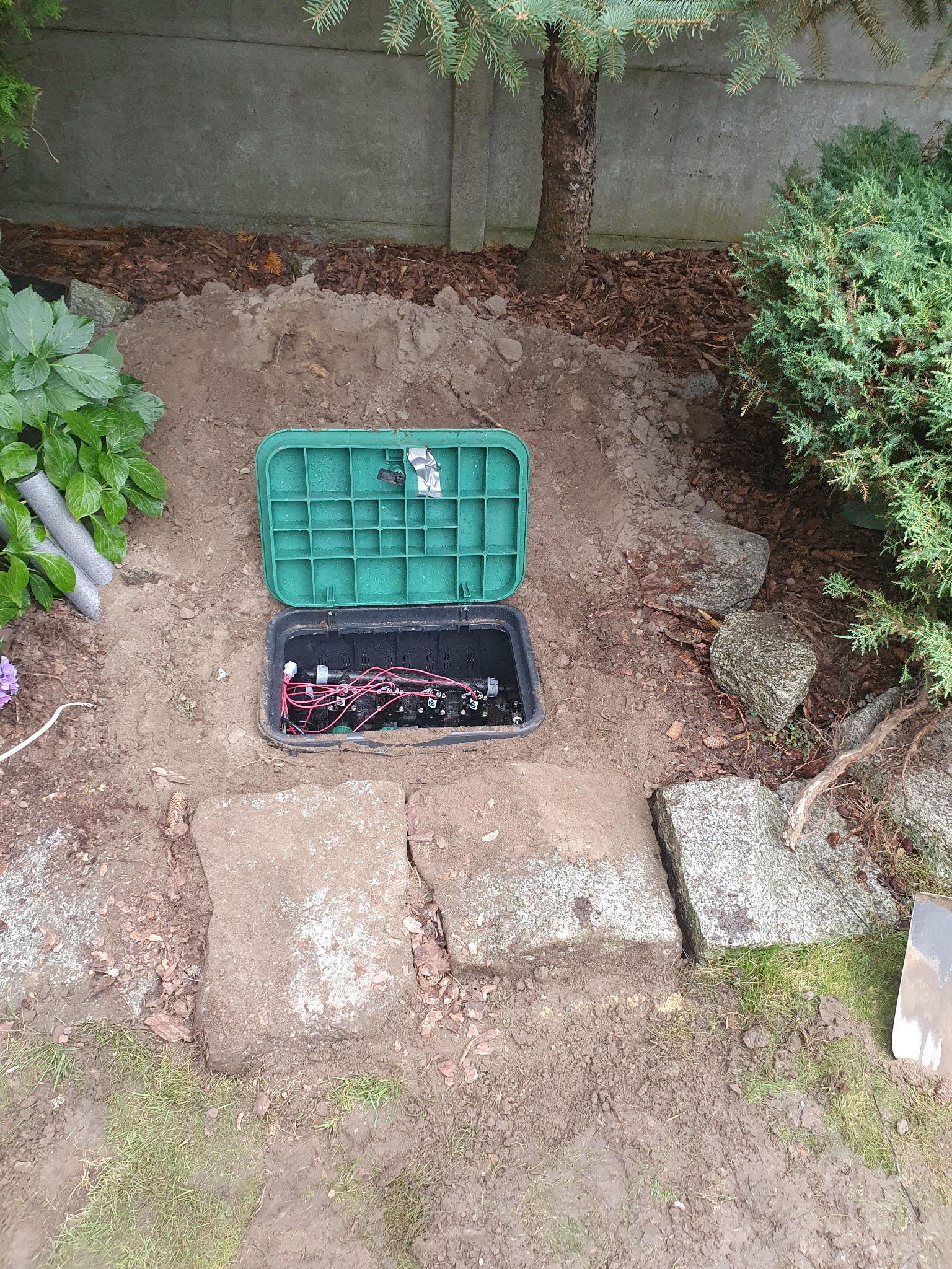 Otwarta, zielona skrzynka sterująca systemem nawadniania w wykopie, widoczne przewody i złączki, otoczenie z ziemi, kamieni i roślinności.