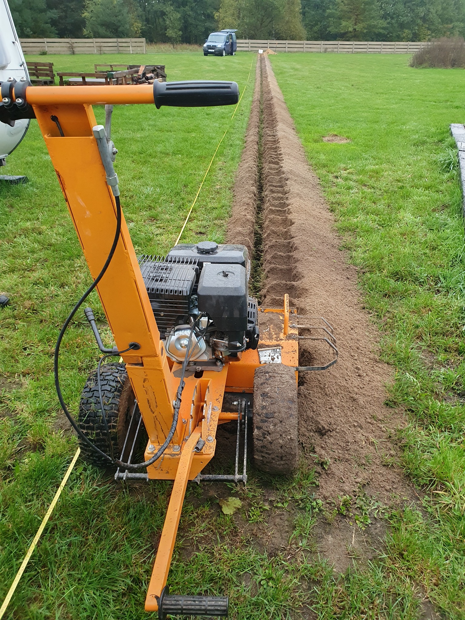 Pomarańczowa glebogryzarka spalinowa tworzy wąski wykop w trawniku, widoczna linia wyznaczająca trasę, w tle ogrodzenie i samochód.