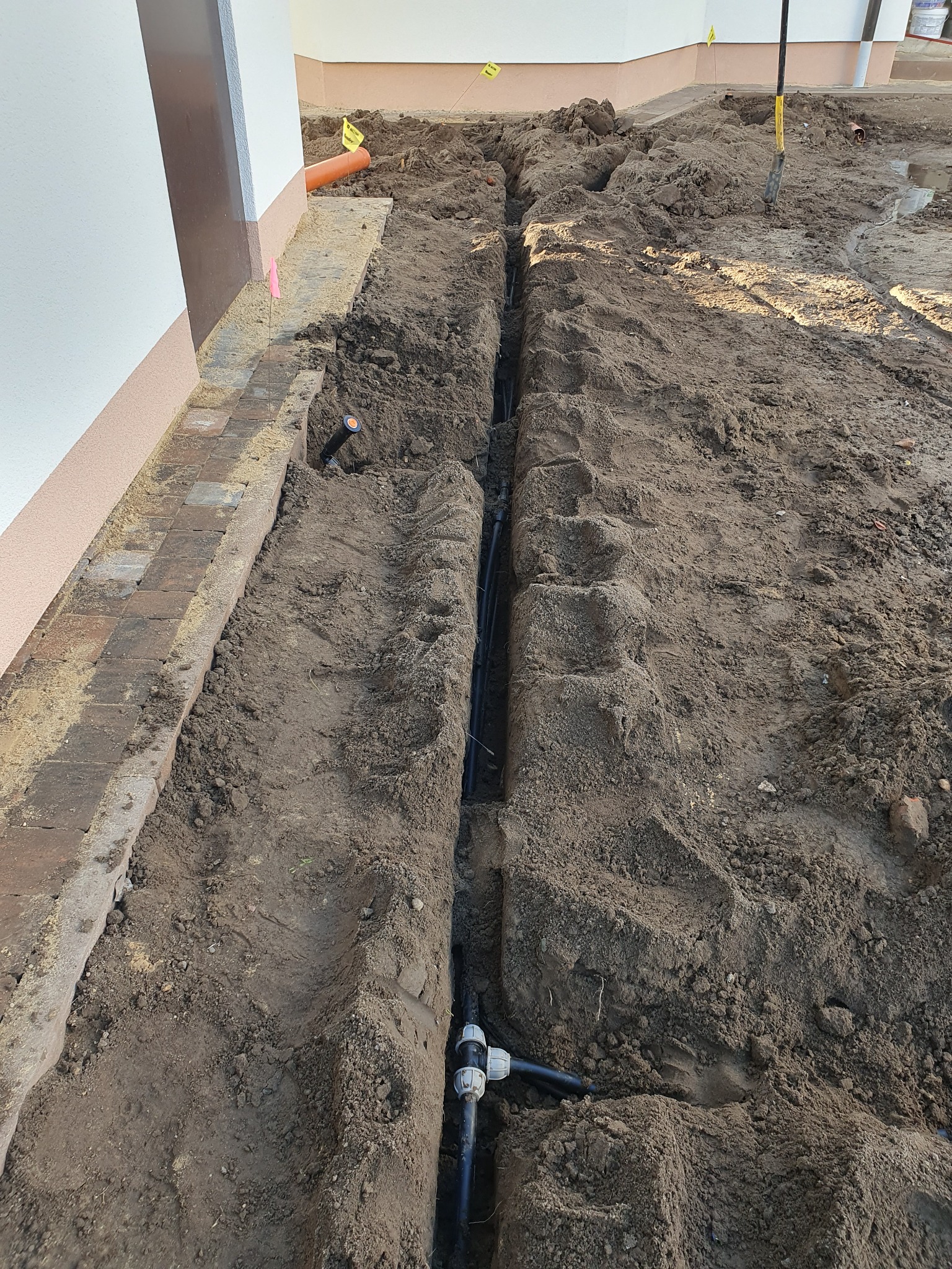 Wykopany rów w ziemi wzdłuż budynku z widocznymi czarnymi rurami instalacyjnymi i elementami złącznymi, częściowo zasypany ziemią, z fragmentem pomarańczowej rury wystającej z ziemi w tle.