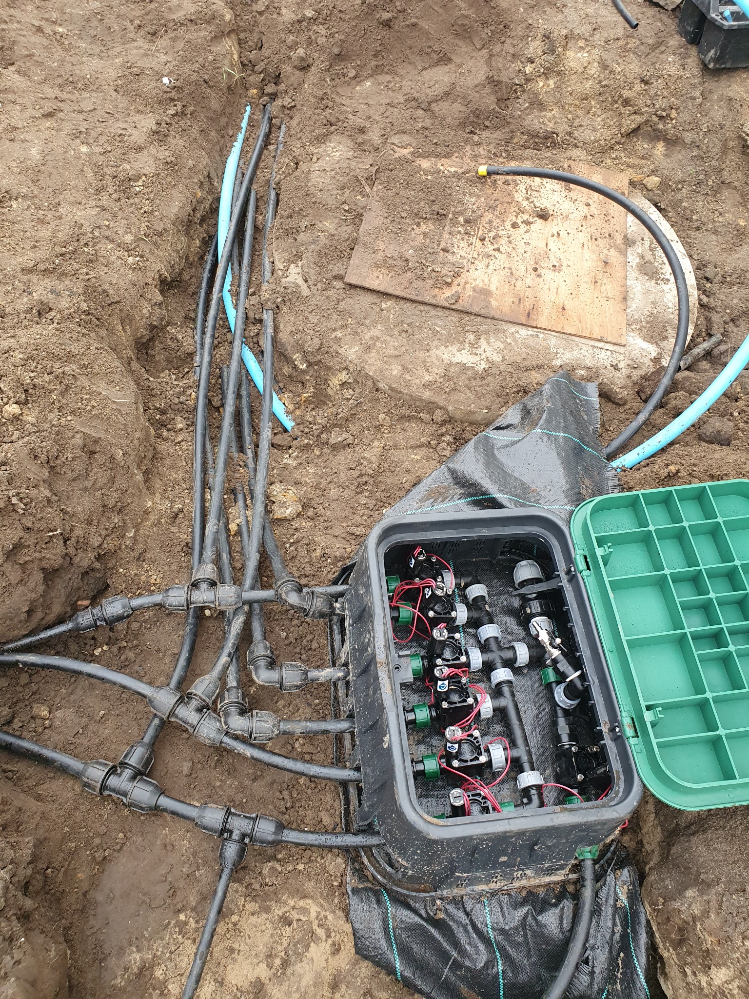 Podziemna instalacja nawadniająca w trakcie montażu: widoczna skrzynka sterująca z elektrozaworami, rury doprowadzające wodę oraz czarna agrowłóknina. Świeżo wykopany rów z przewodami.