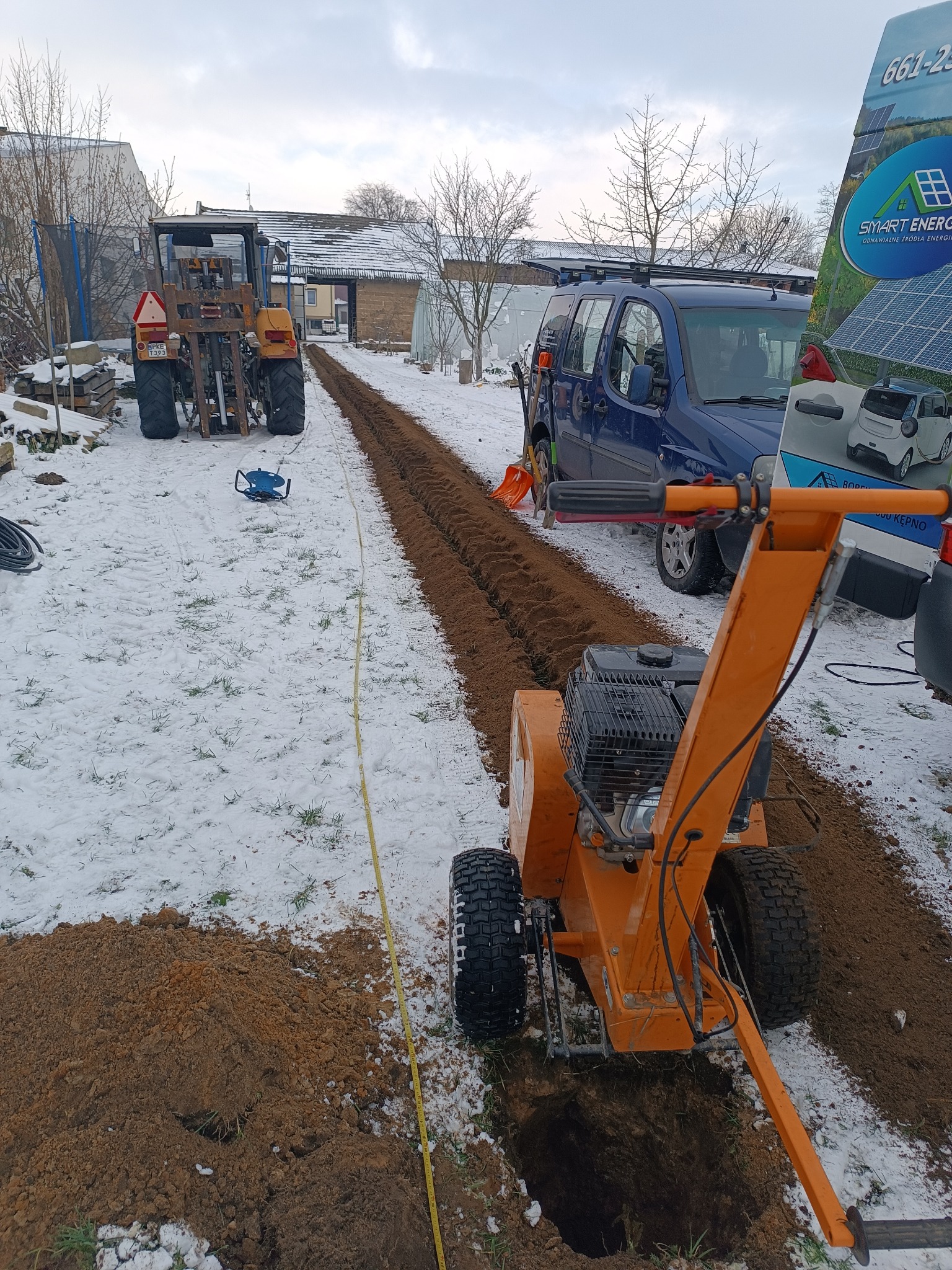 Pomarańczowa glebogryzarka tworzy wąski wykop w ziemi pokrytej śniegiem, w tle widoczny traktor, samochód dostawczy i reklama paneli słonecznych.