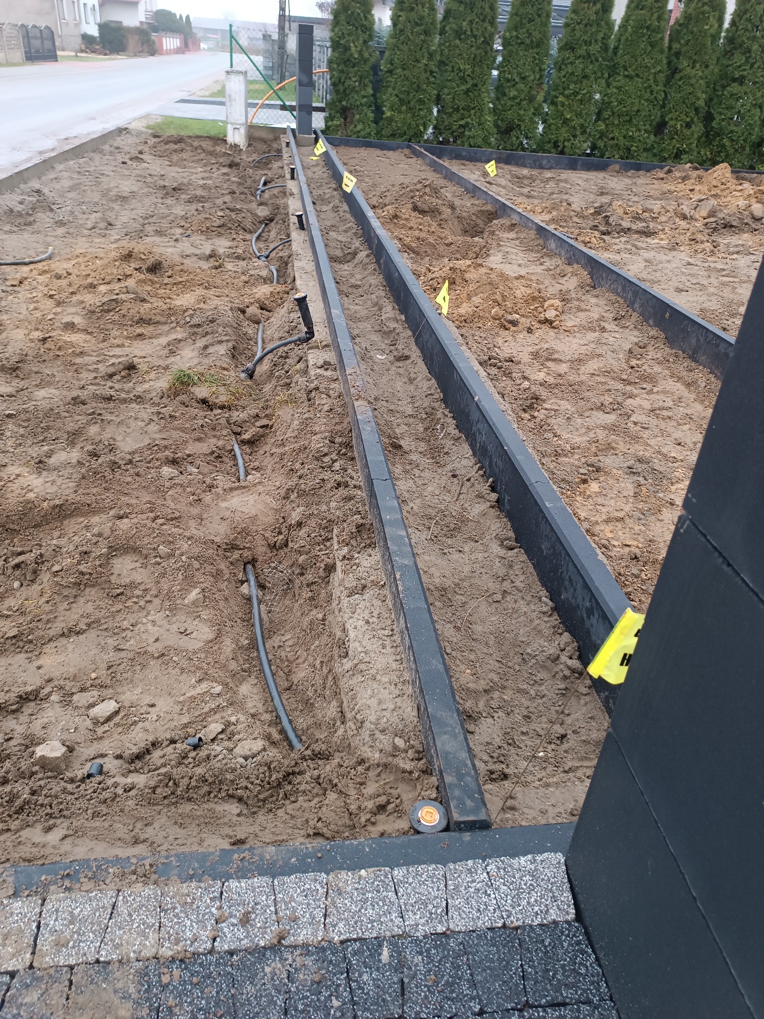 Przygotowanie terenu pod system nawadniania: wykopane rowy z czarnymi obrzeżami, widoczne rury nawadniające w ziemi, żółte znaczniki w wykopach.