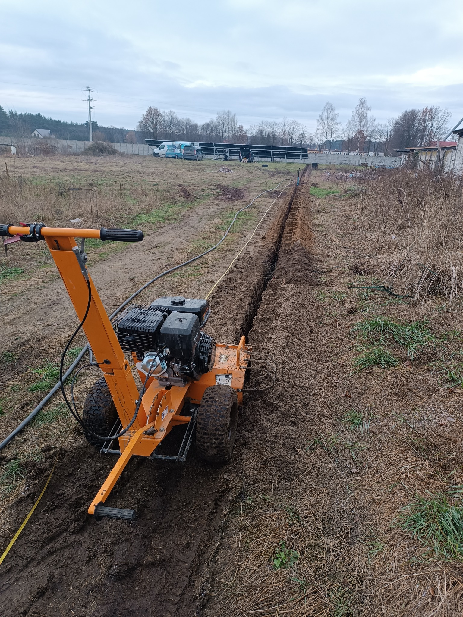 Pomarańczowa glebogryzarka spalinowa tworzy wąski wykop w ziemi na zewnątrz, widoczna linia wykopu ciągnąca się w dal, w tle zabudowania i pochmurne niebo.
