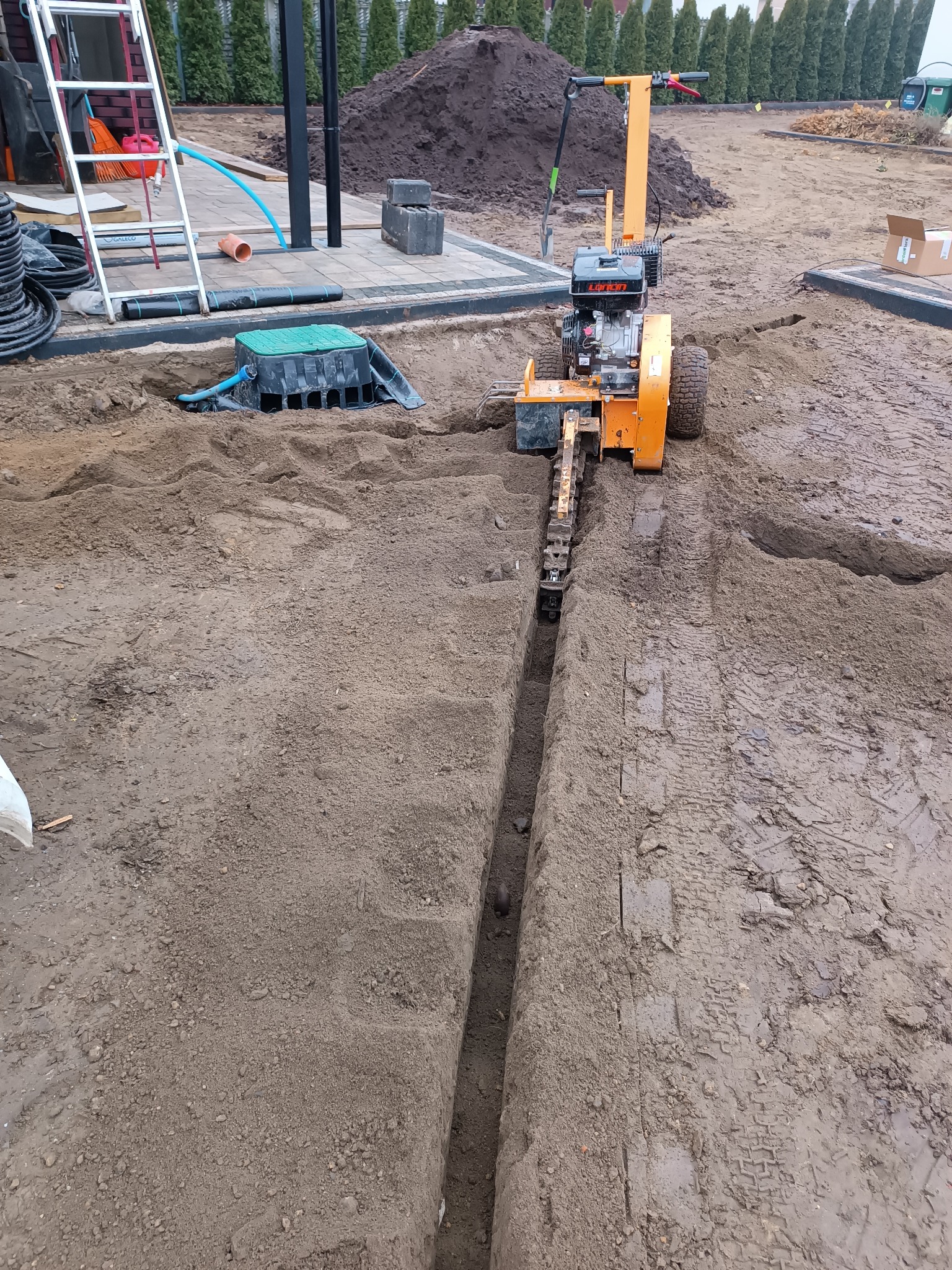 Pomarańczowa glebogryzarka wykopuje wąski rów w ziemi, widoczny fragment instalacji podziemnej, na drugim planie trawnik z tujami.