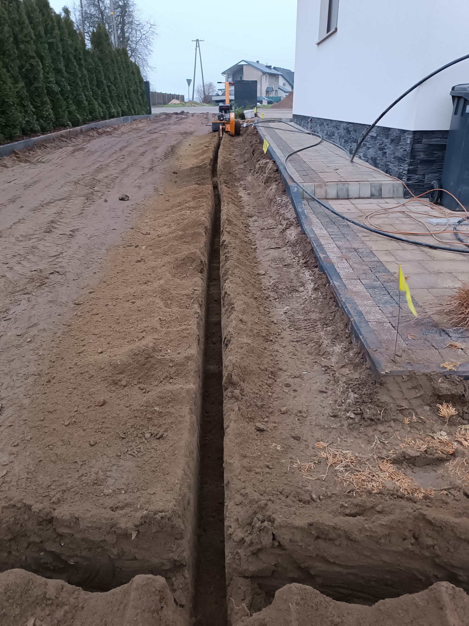 Wąski wykop w ziemi prowadzony wzdłuż chodnika z kostki brukowej przy budynku mieszkalnym, z żółtą flagą sygnalizacyjną i mini koparką w tle.