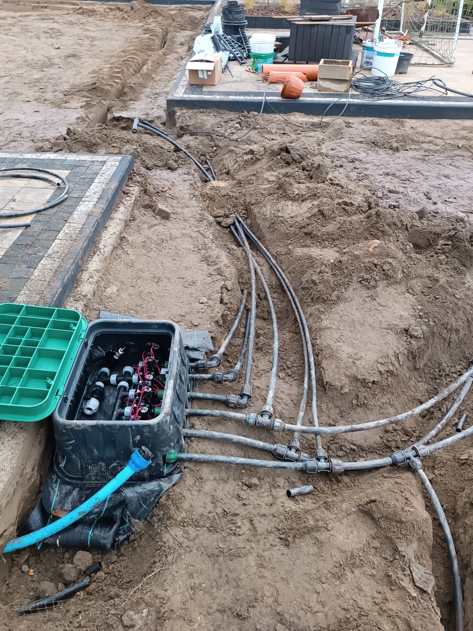 Instalacja podziemnego systemu nawadniania: skrzynka sterująca z otwartą zieloną pokrywą, rury rozprowadzające w wykopie, ziemia wokół.