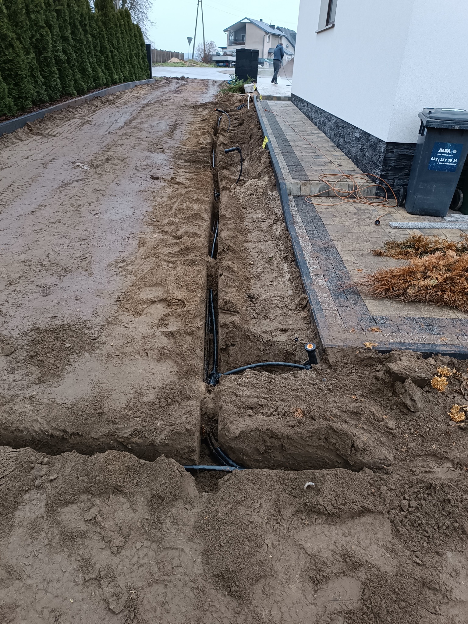 Wykopany rów na posesji z widocznymi rurami instalacji nawadniającej, częściowo zasypany ziemią, obok kostka brukowa i śmietnik.