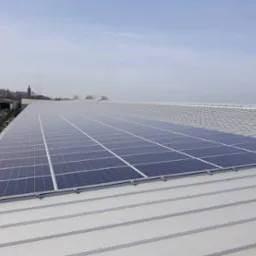 Rząd paneli słonecznych na metalowym dachu budynku przemysłowego, widok z góry, w tle zabudowania i kościół.