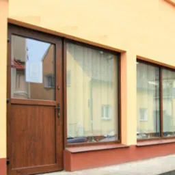 Fasada budynku z brązowymi oknami PCV i drzwiami. Widoczne rolety wewnętrzne w oknach, odbicie budynku naprzeciwko. Jasna elewacja z ciemniejszą podmurówką.