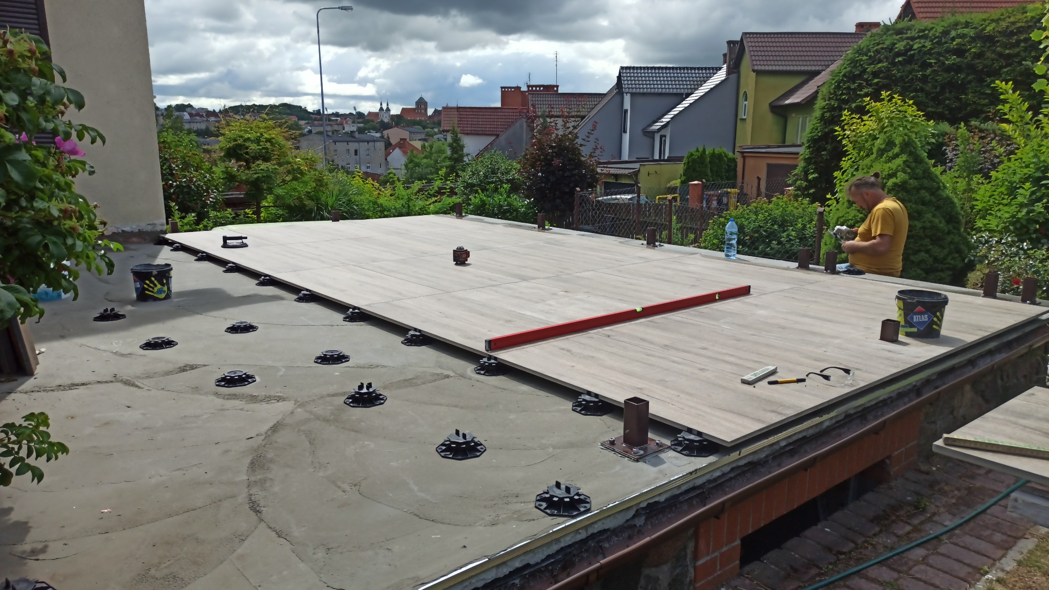 Montaż desek tarasowych na regulowanych wspornikach na dachu, poziomica i laser krzyżowy na powierzchni, pracownik w żółtej koszulce przy pracy, widok na miasto w tle.