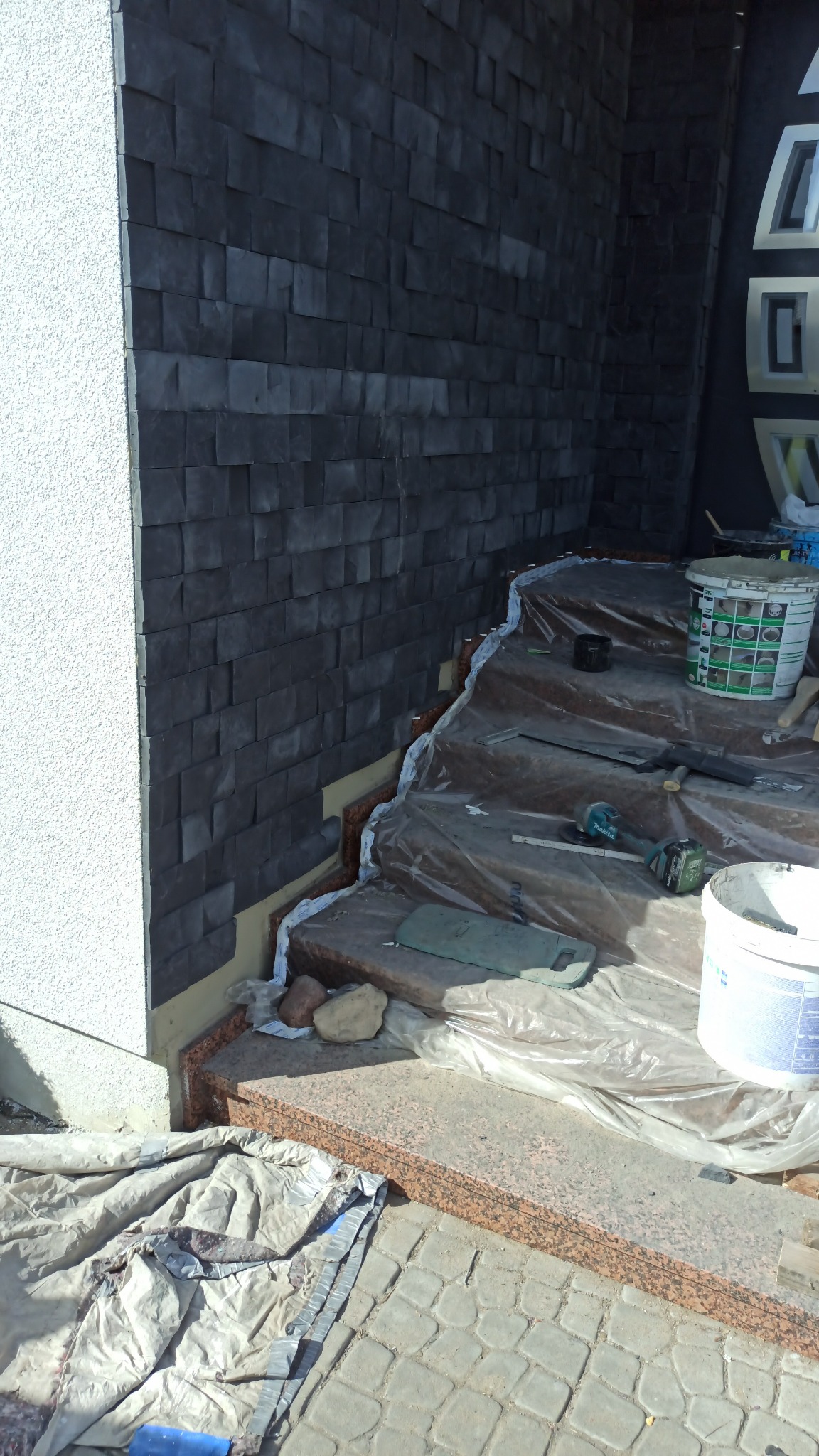 Montaż okładziny z ciemnego kamienia elewacyjnego na ścianie przy schodach wejściowych, zabezpieczonych folią malarską, widoczne narzędzia i wiadro z zaprawą.