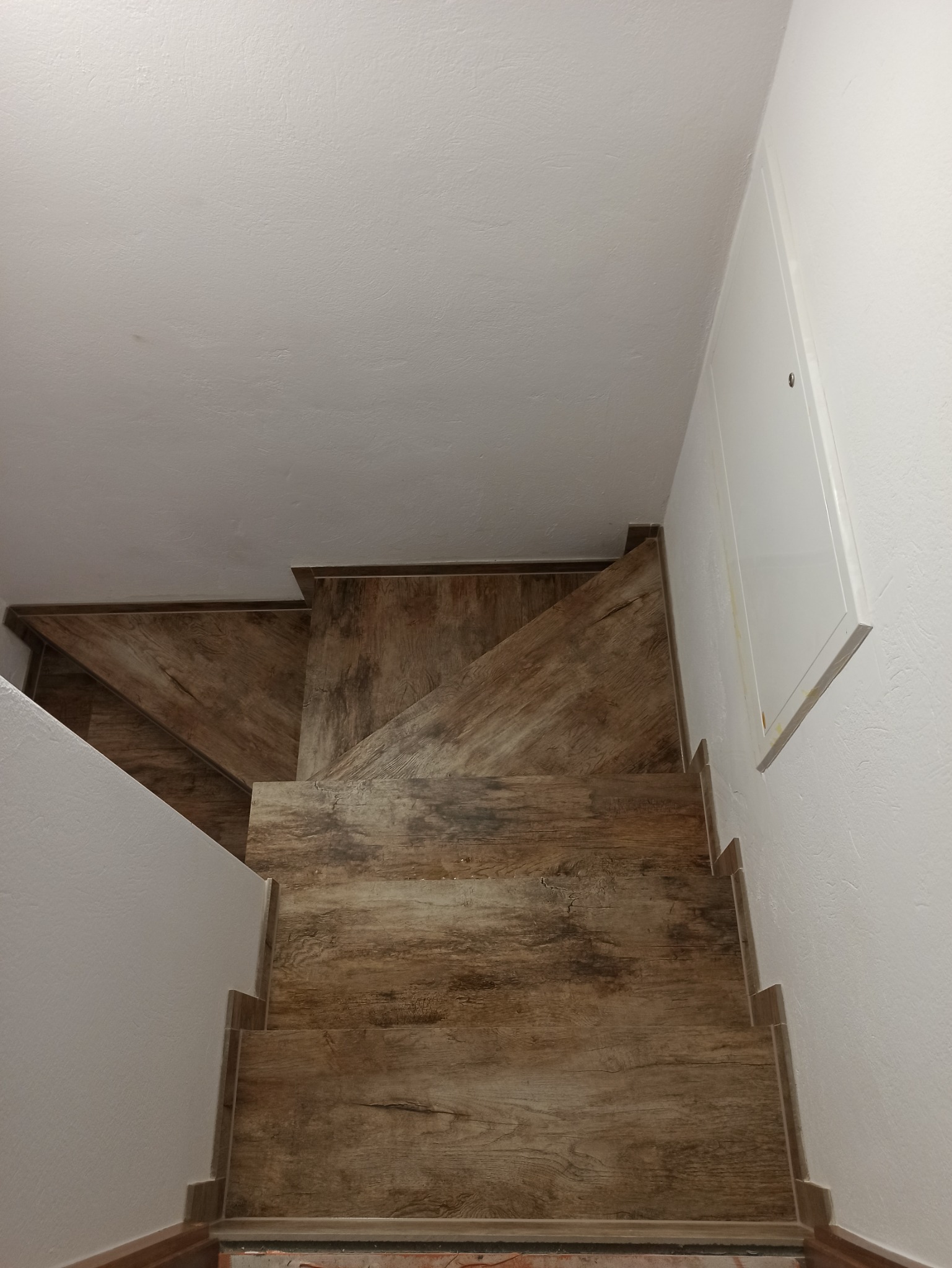 Schody wyłożone panelami imitującymi drewno, widok z góry, białe ściany i białe drzwi rewizyjne z boku.