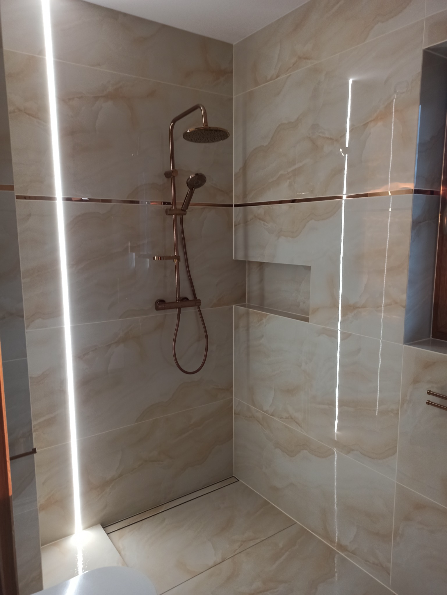 Nowoczesna kabina prysznicowa z deszczownicą w kolorze miedzi, wykończona płytkami imitującymi marmur z dekoracyjną, miedzianą listwą i wnęką w ścianie.