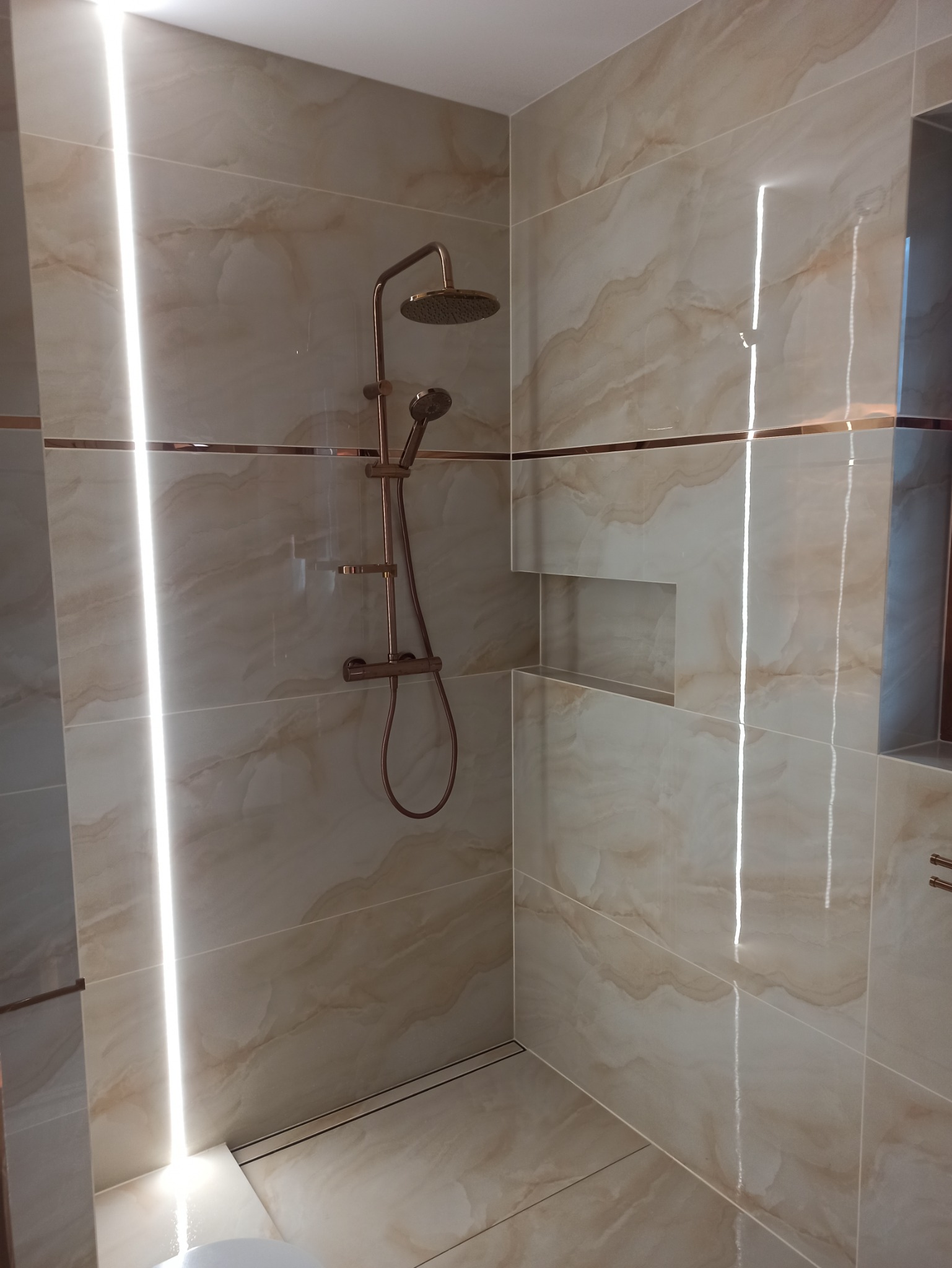 Wykończona łazienka z beżowymi płytkami imitującymi marmur, z wnęką w ścianie i deszczownicą w kolorze różowego złota.