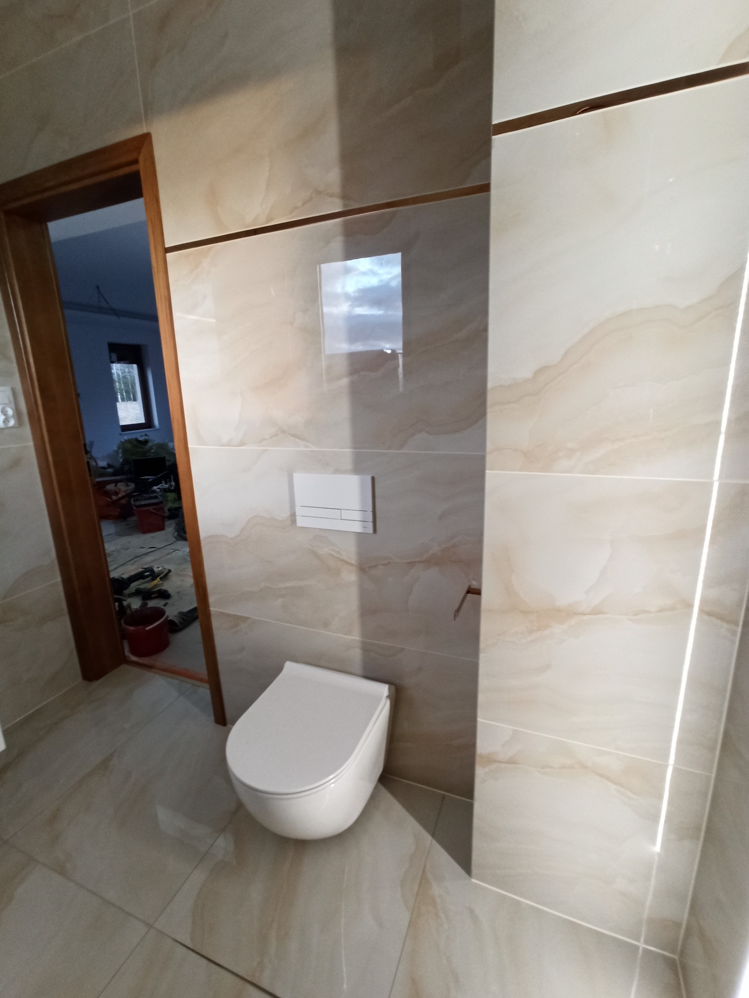 Nowoczesna łazienka z beżowymi płytkami imitującymi marmur, podwieszana toaleta i widoczne narzędzia remontowe w tle, sugerujące trwające prace wykończeniowe.