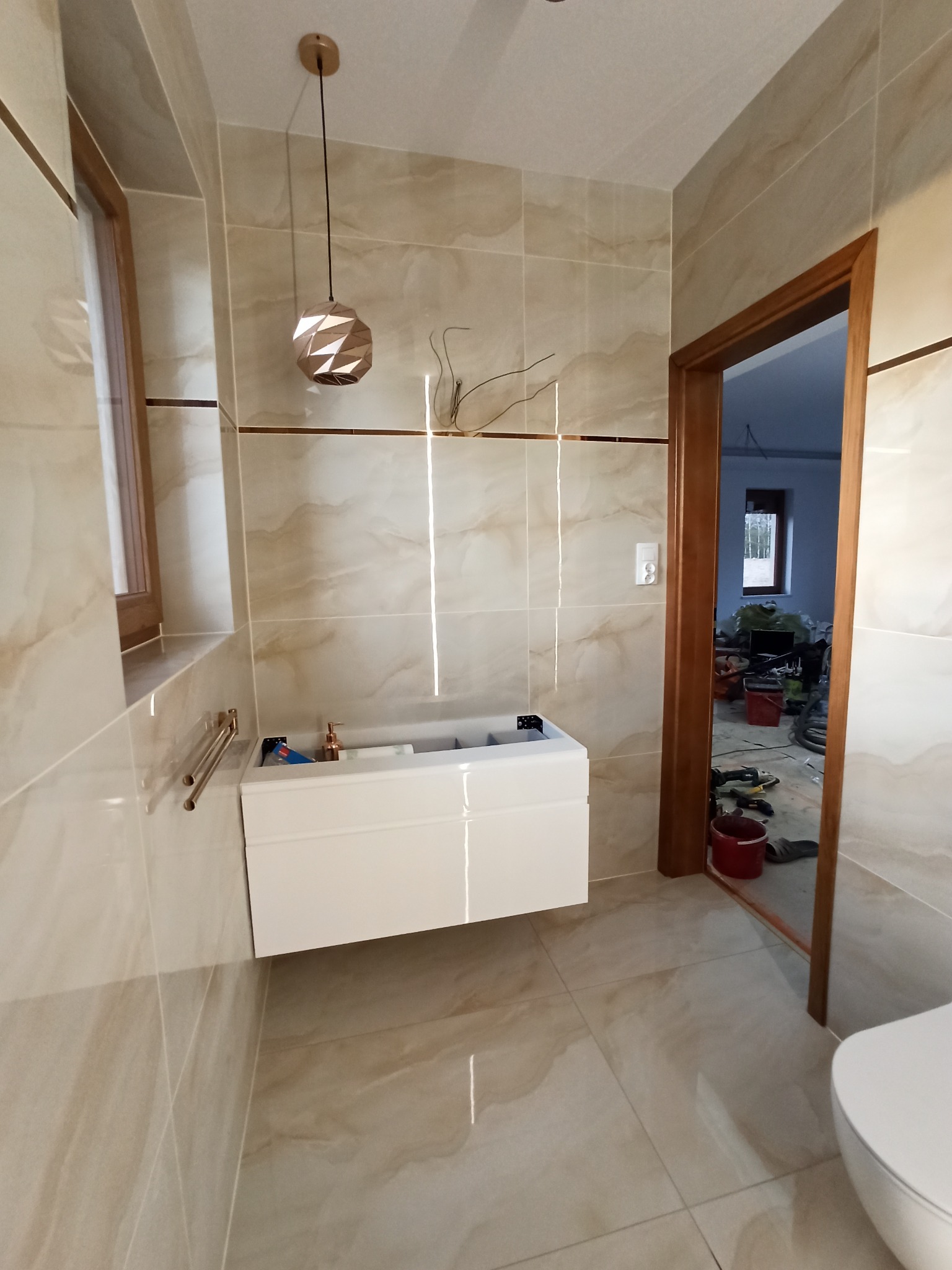 Łazienka w trakcie wykańczania, beżowe płytki imitujące marmur, biała szafka pod umywalkę bez blatu, widoczne narzędzia i bałagan w pomieszczeniu obok.