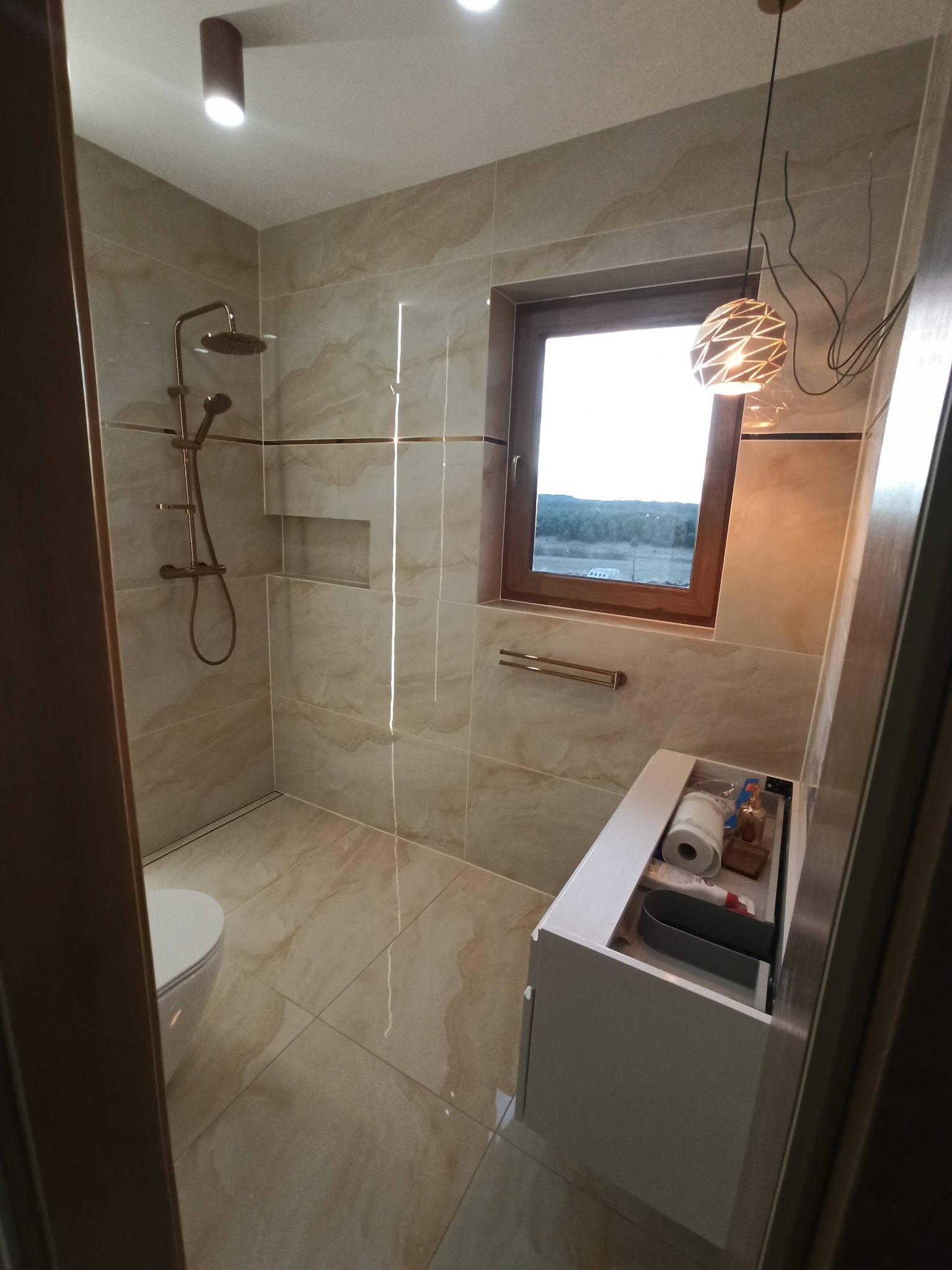 Jasna łazienka z beżowymi płytkami imitującymi marmur, kabina prysznicowa bez brodzika, okno z drewnianą ramą i wisząca lampa z geometrycznym kloszem.