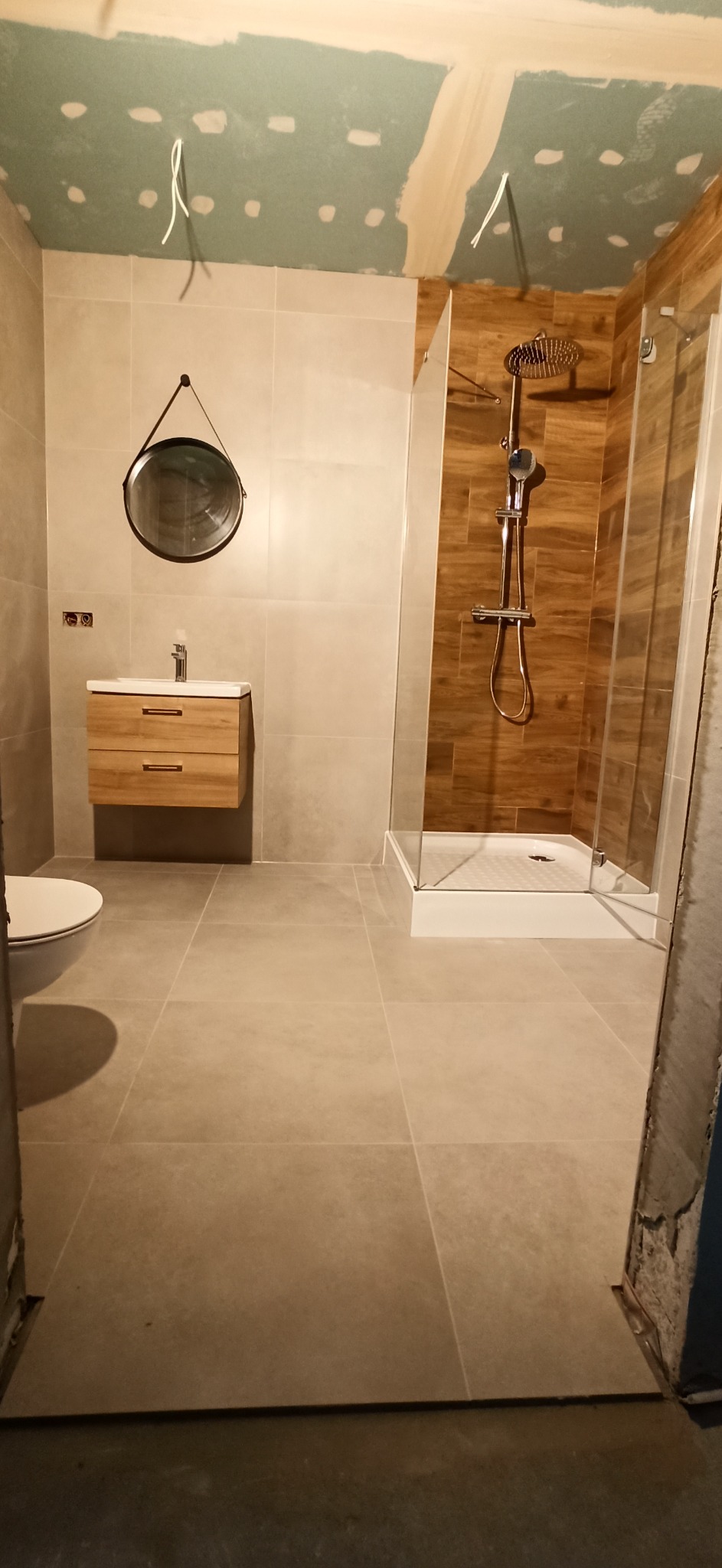 Wnętrze łazienki w trakcie wykańczania, widoczna kabina prysznicowa z drewnopodobnymi płytkami, umywalka z szafką z jasnego drewna, okrągłe lustro z czarną ramą oraz częściowo wykończone ściany...