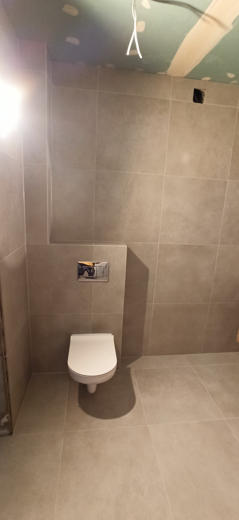 Niedokończona łazienka z szarymi płytkami, wiszącą toaletą i wystającymi kablami z sufitu.