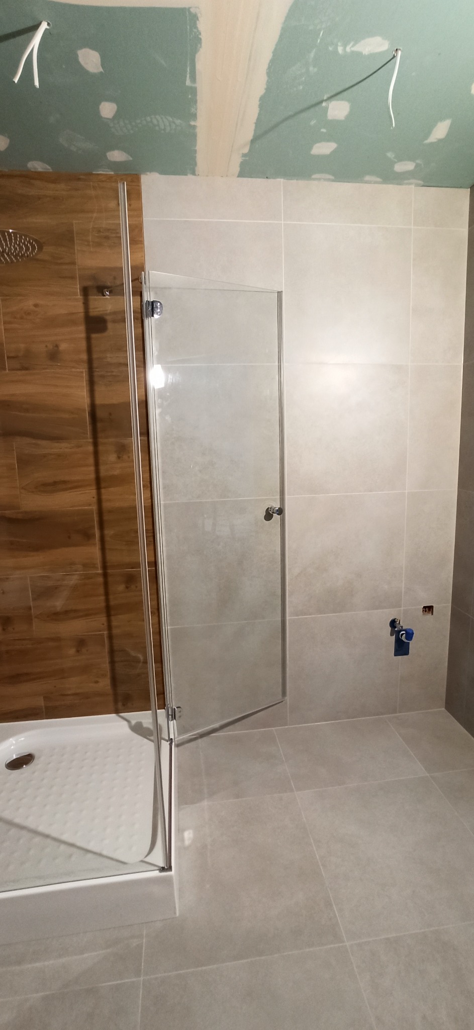 Niedokończona łazienka z zamontowaną kabiną prysznicową z drewnopodobnymi i beżowymi płytkami, widoczne niedoskonałości na suficie i wystające kable.